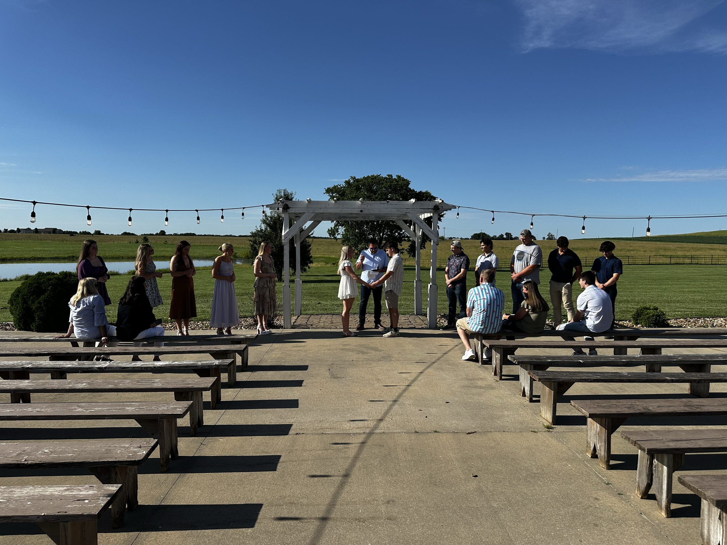 A group of people gathered outdoors under a white pergola, with benches in the foreground and a blue sky overhead, on a sunny day in a rural area.
