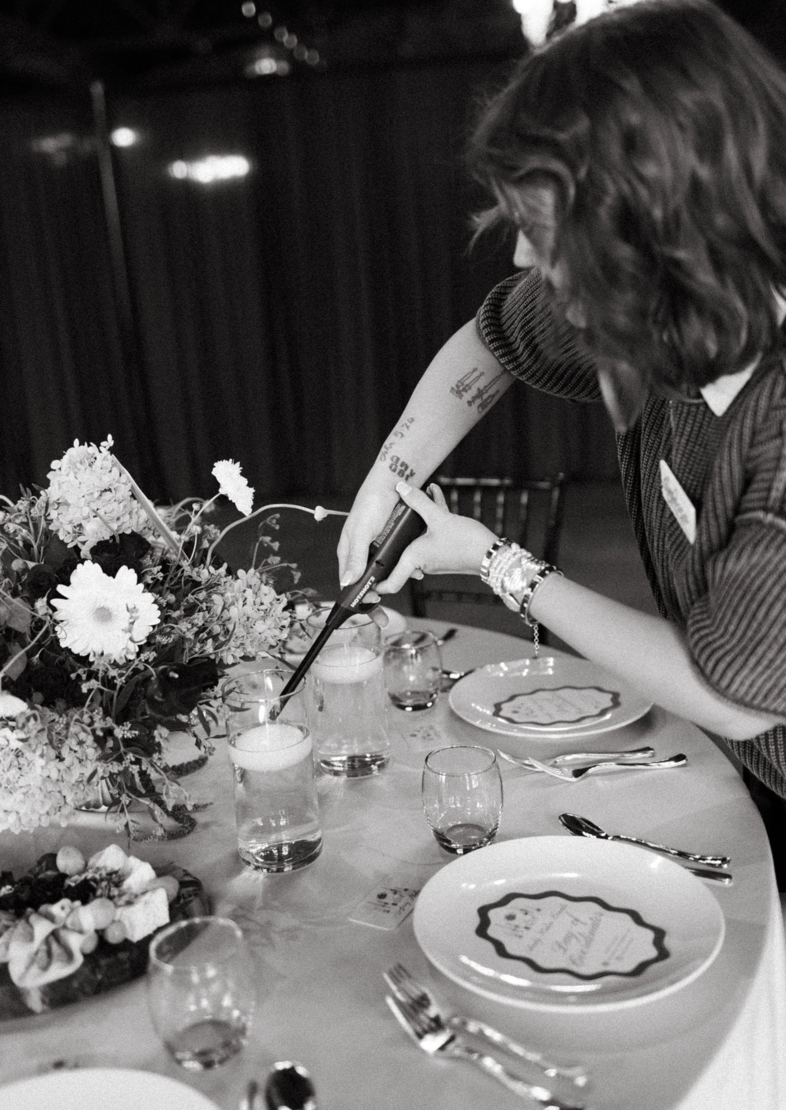 A woman with wavy hair and tattoos on her arm is filling a glass of wine at a dinner table with floral arrangements, plates, silverware, and glasses.