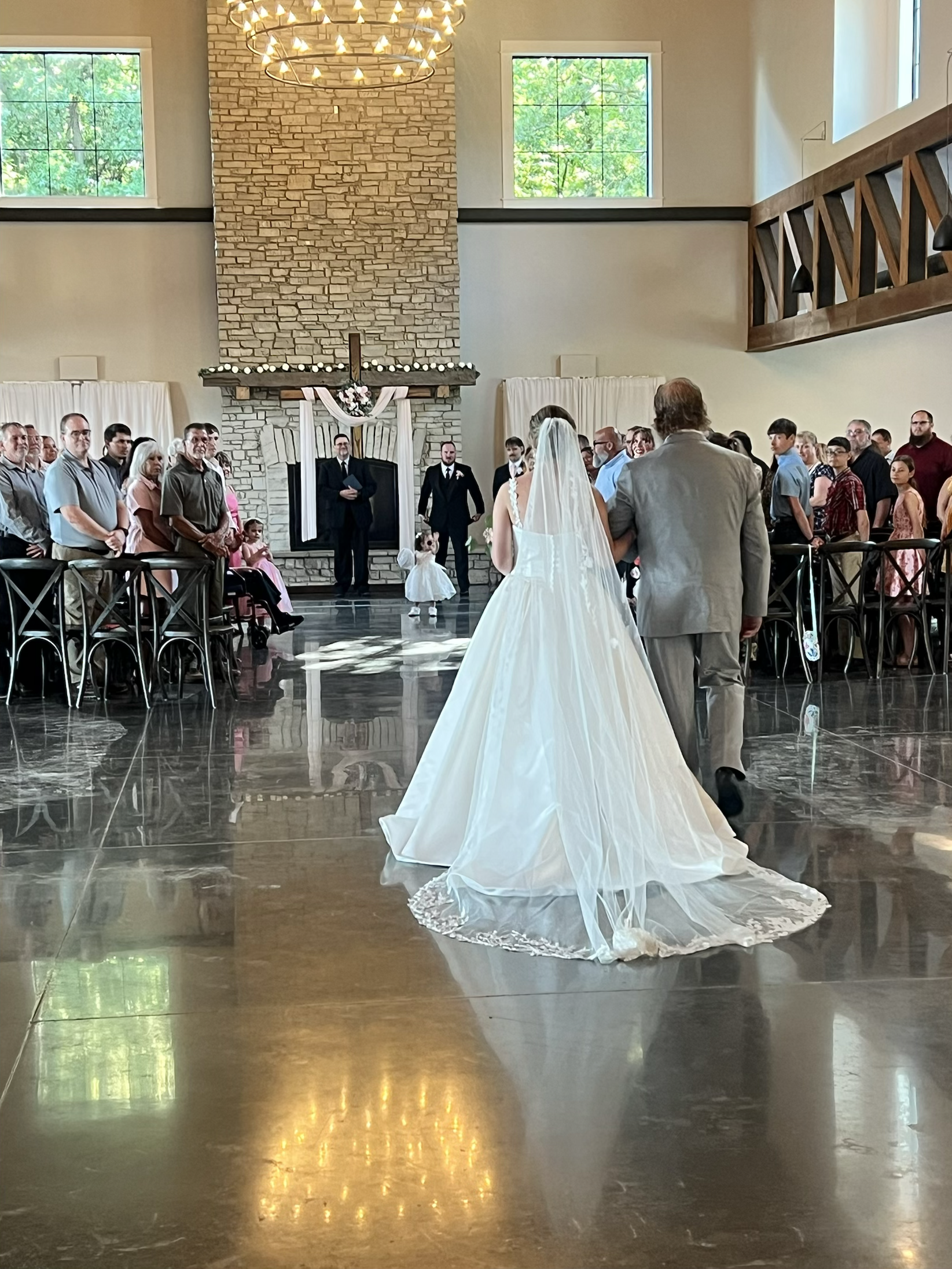 Bride and groom standing before officiant during wedding ceremony inside a church with large brick fireplace, high windows, and decor including ribbons and flowers.