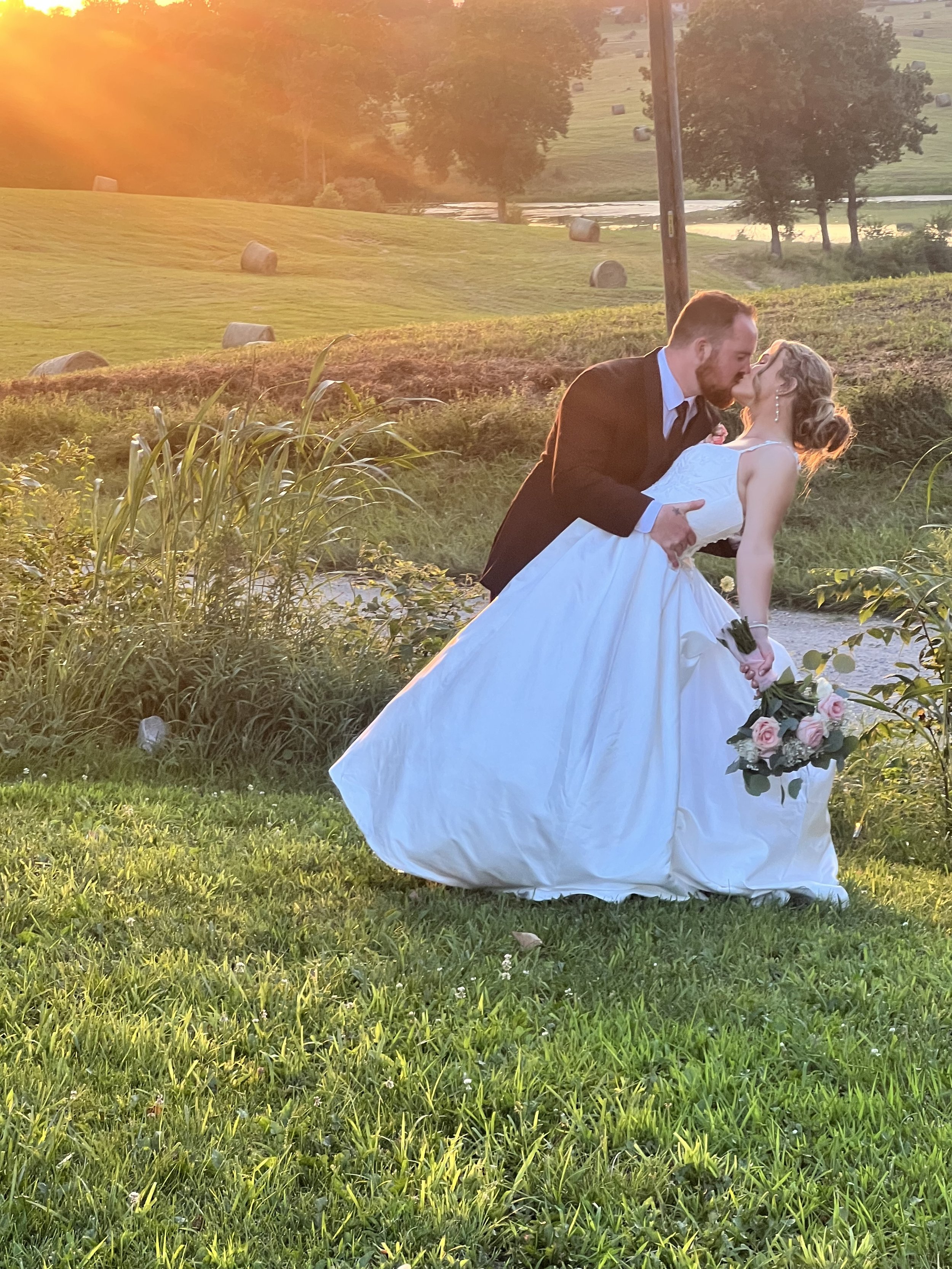 A newlywed couple sharing a kiss outdoors during sunset, with the bride holding a bouquet of pink and white roses, in a lush green landscape with hay bales and trees in the background.