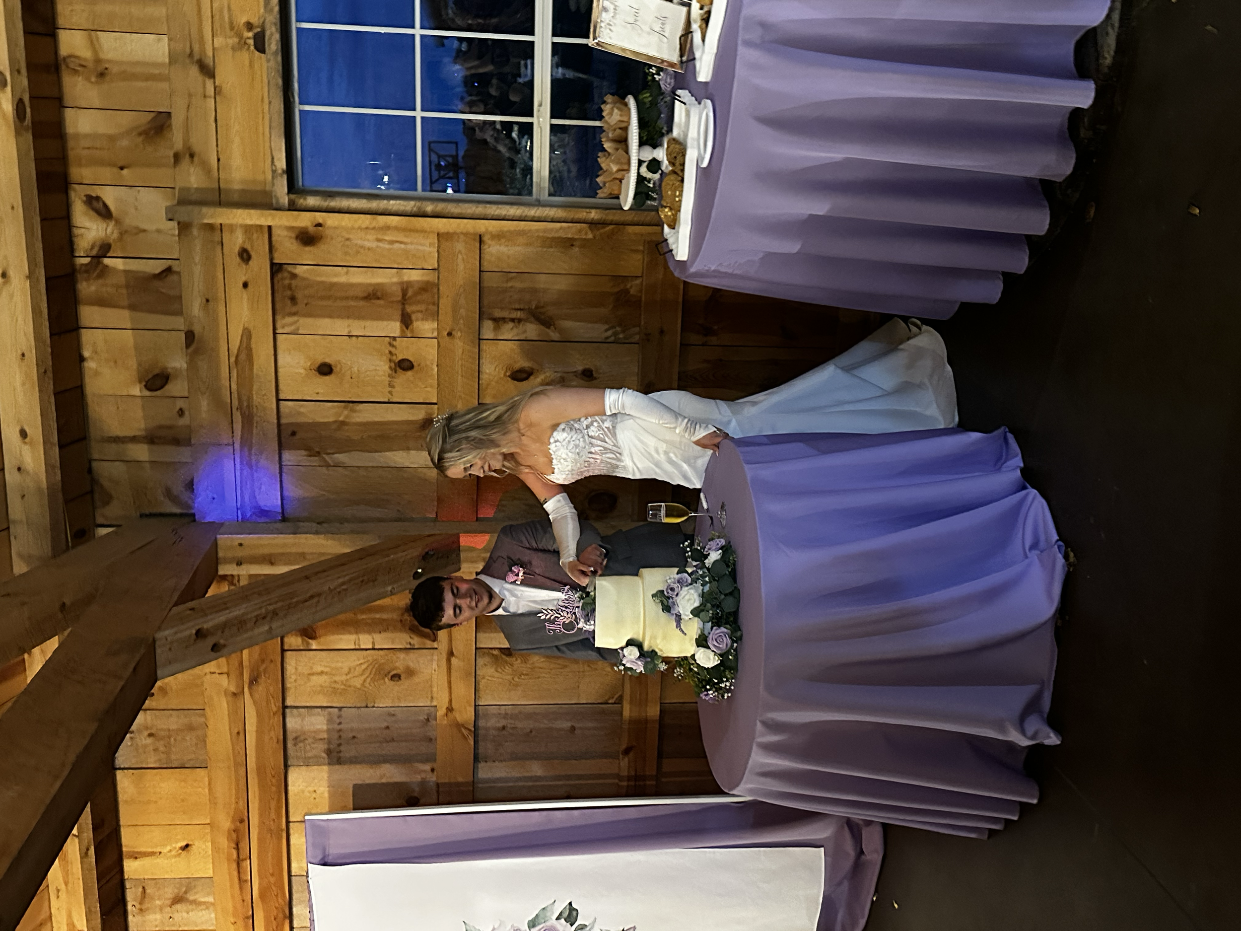 Bride in white wedding dress and groom in gray suit standing behind a wedding cake at reception with purple tablecloths and wooden wall background.