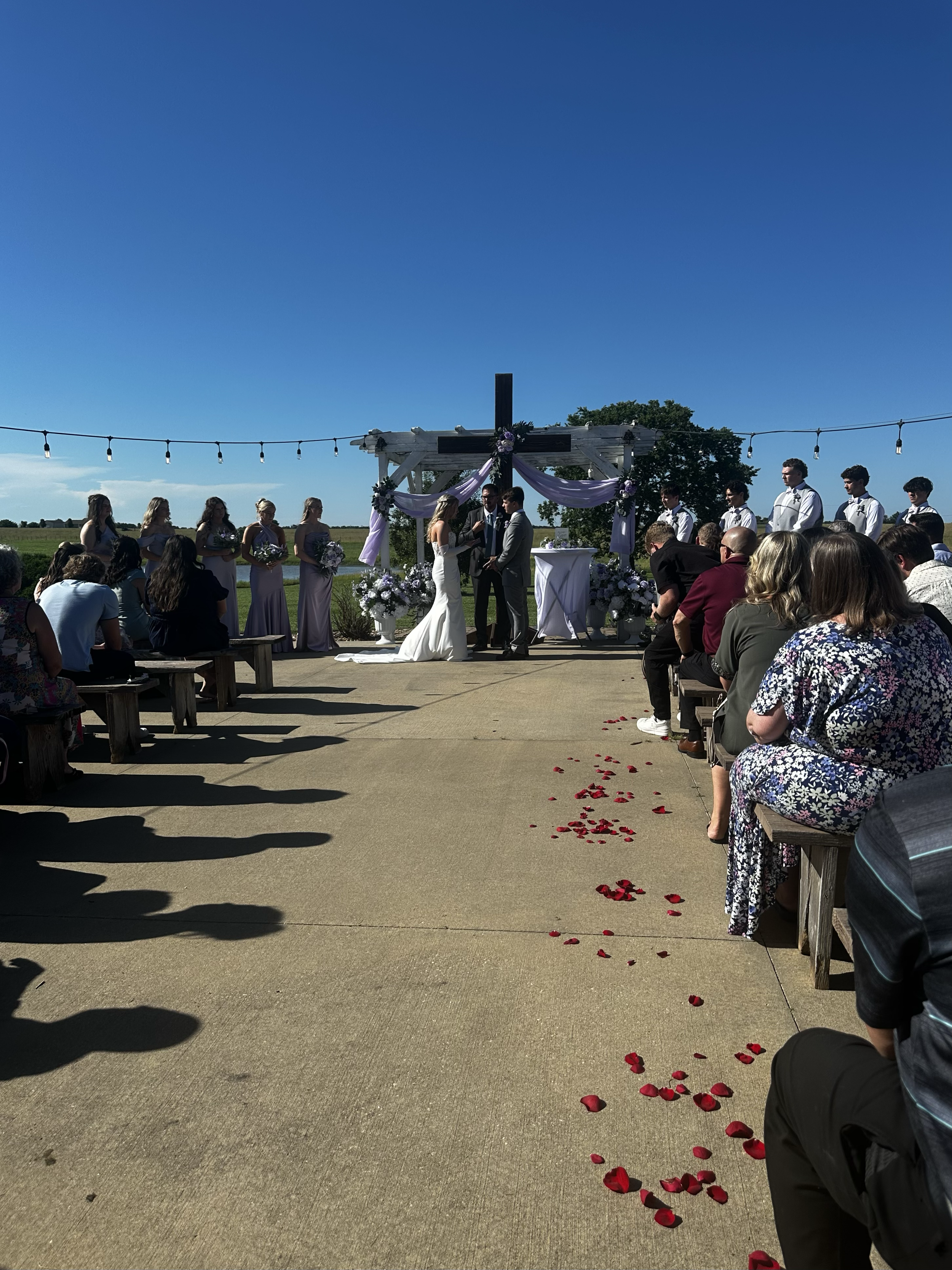 Outdoor wedding ceremony with a cross decorated with purple and white fabric, surrounded by flower arrangements, under string lights, with guests seated on benches, and a clear blue sky.