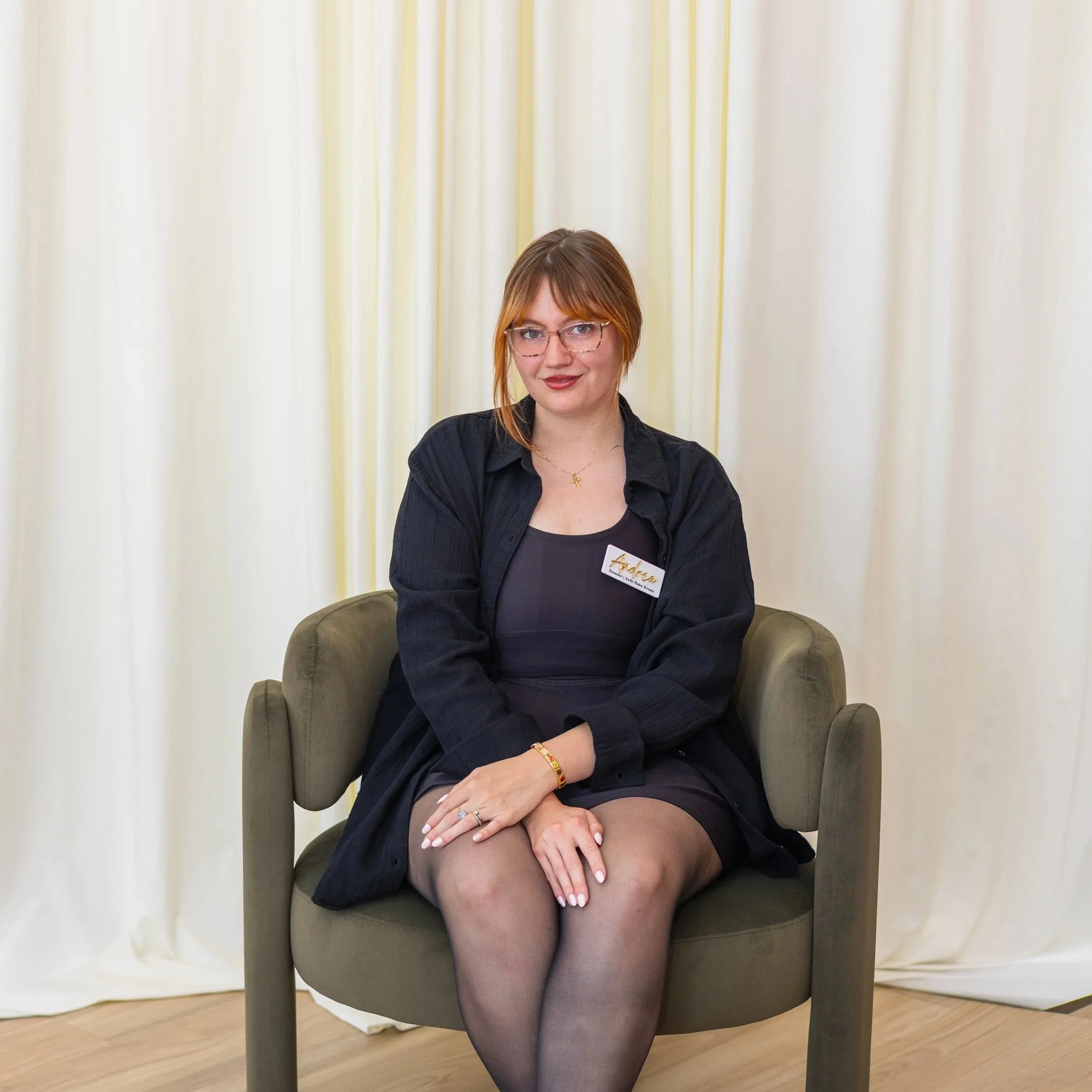 A woman with red hair, glasses, and a nose piercing sitting with her legs crossed, wearing a black shirt and sheer tights, in front of a white curtain