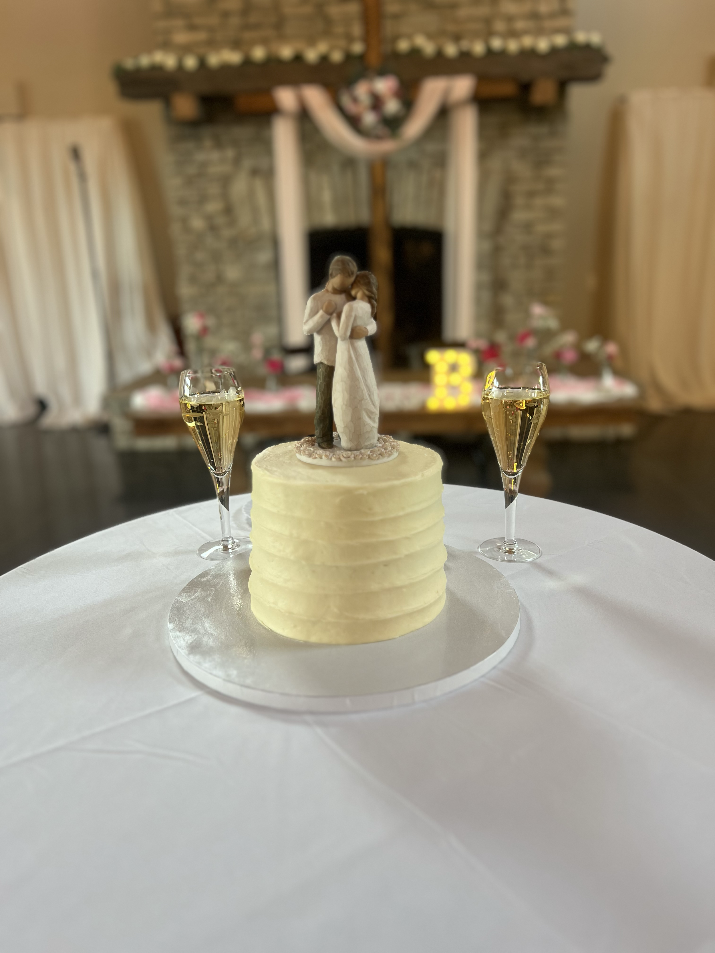 A wedding cake with a bride and groom figurine topper, two glasses of champagne, and a blurred background of a fireplace and floral decorations.