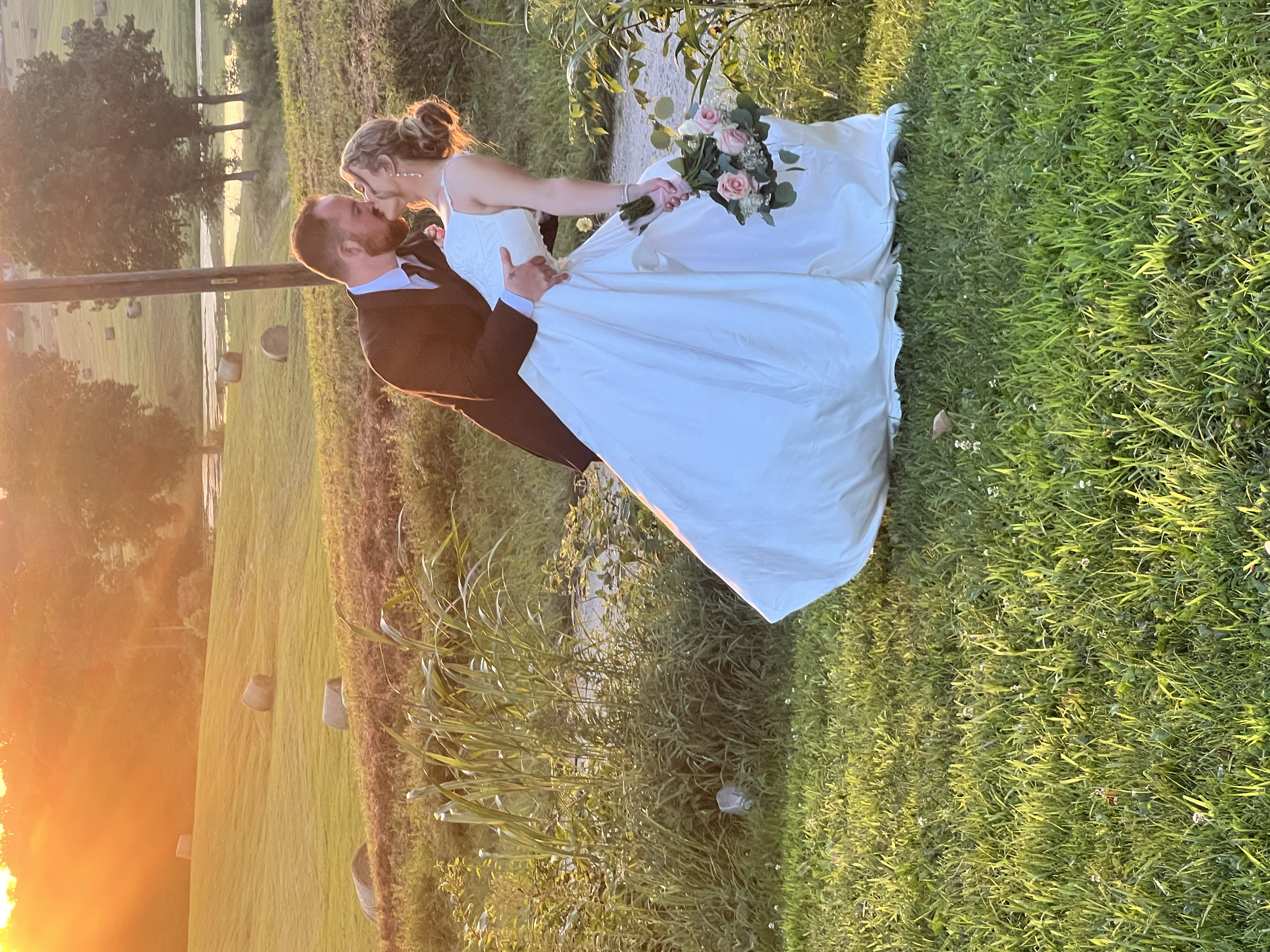 A bride and groom share a kiss outdoors during a sunset, with the bride holding a bouquet of pink and white roses. 