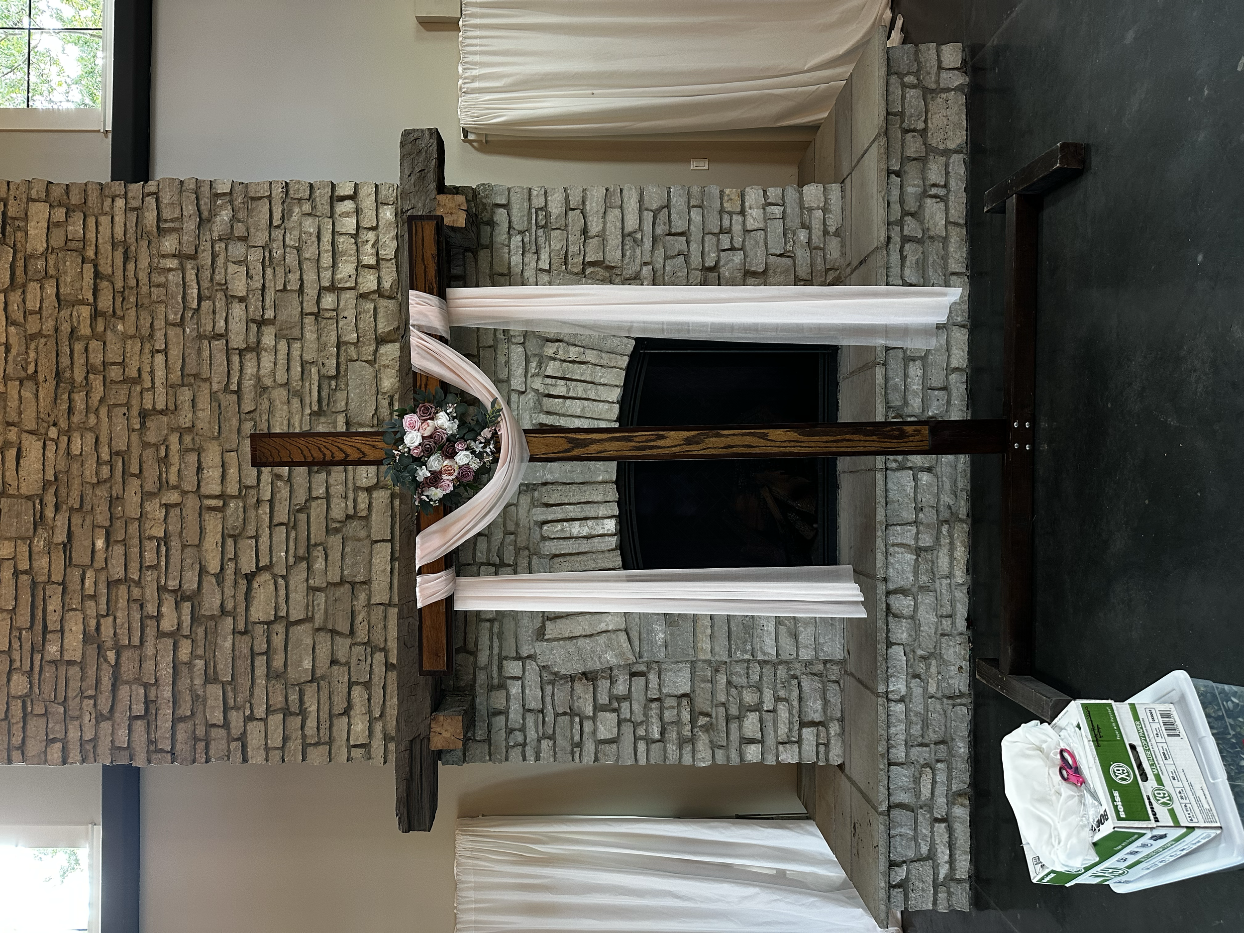 Interior of a room with a brick fireplace, decorated with a pink and white floral arrangement, and sheer curtains covering a window.