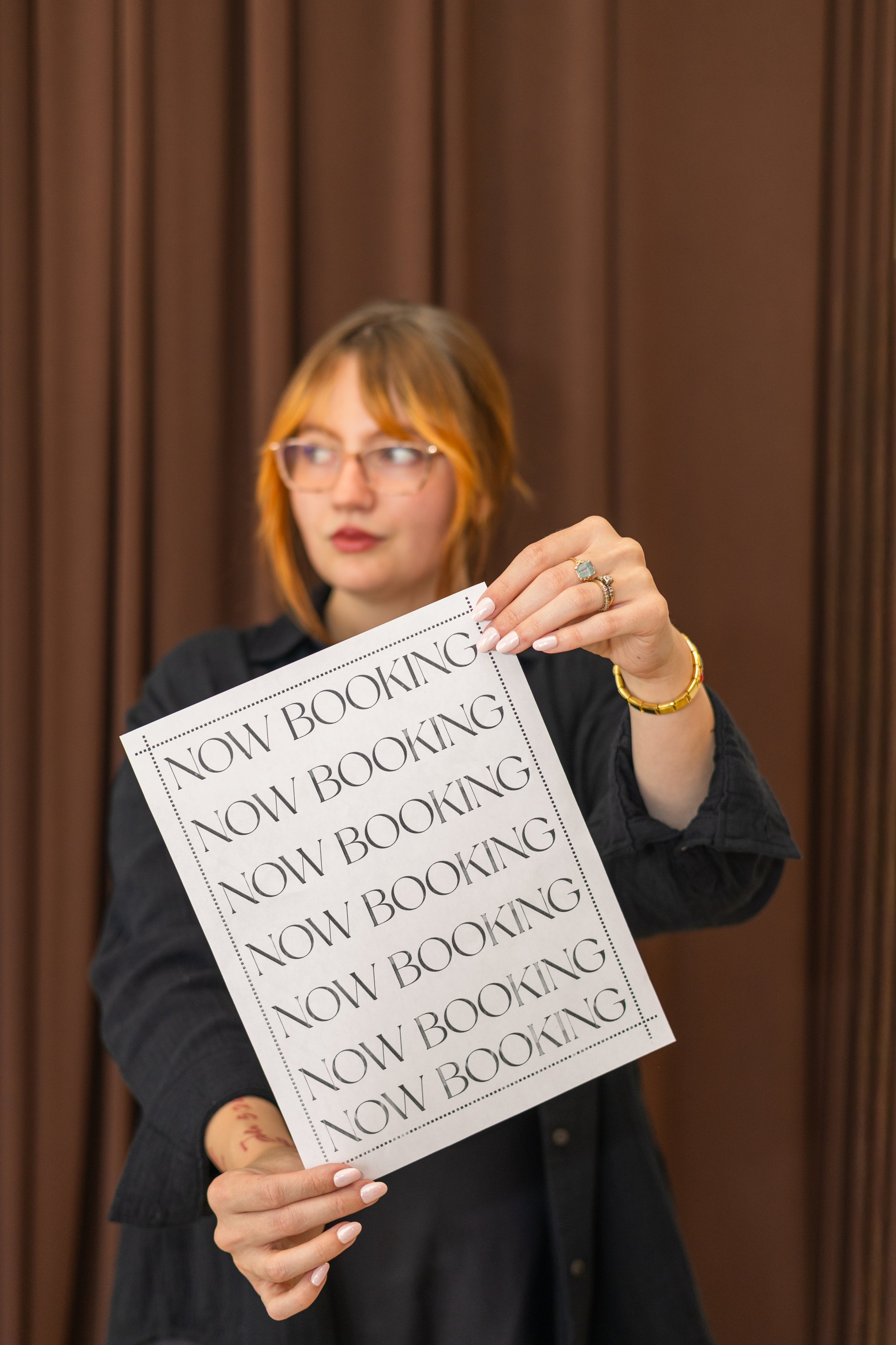 A woman with red hair and glasses holding a paper that says 'NOW BOOKING' repeatedly, standing in front of brown curtains.