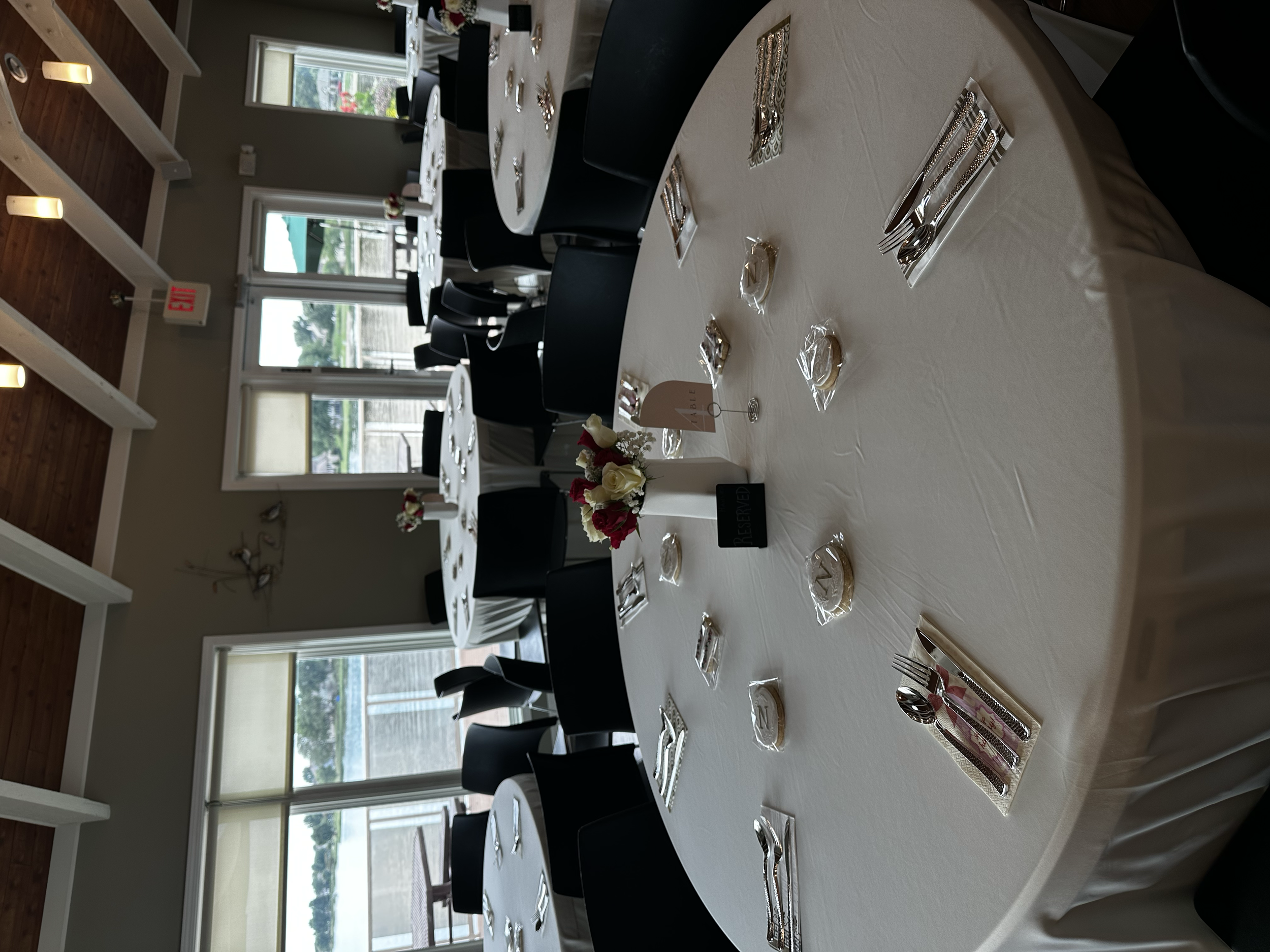 A banquet table setup with white tablecloths, plates, silverware, napkins, small bags, and a flower centerpiece, in a room with large windows and a wooden ceiling.