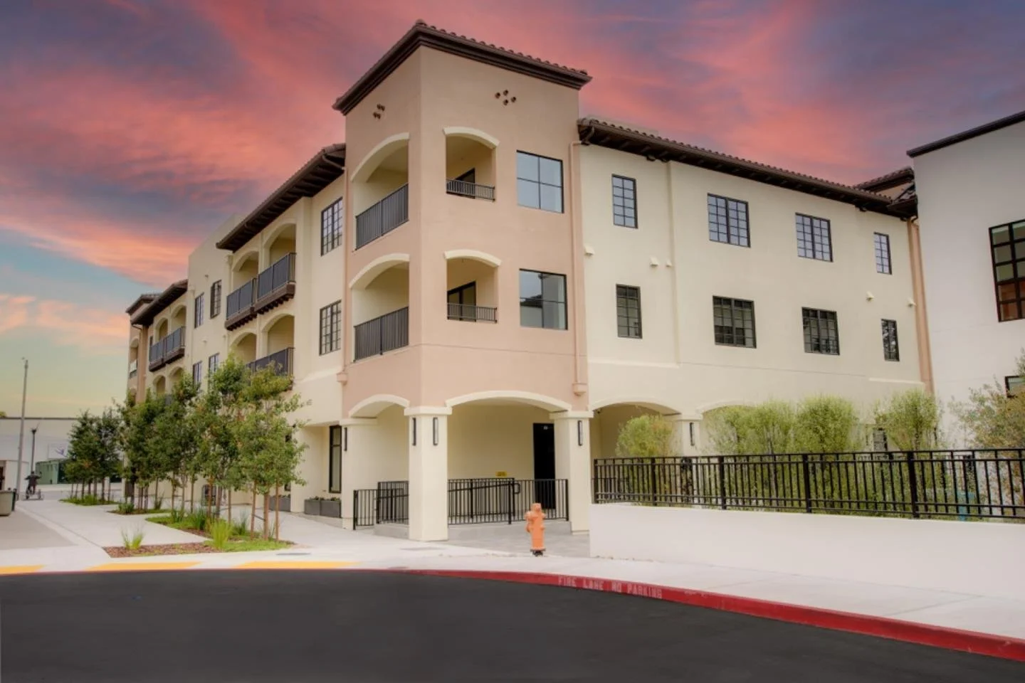 A modern multi-story apartment building with balconies, set against a colorful sunset sky, surrounded by manicured trees and a sidewalk with a fire hydrant.