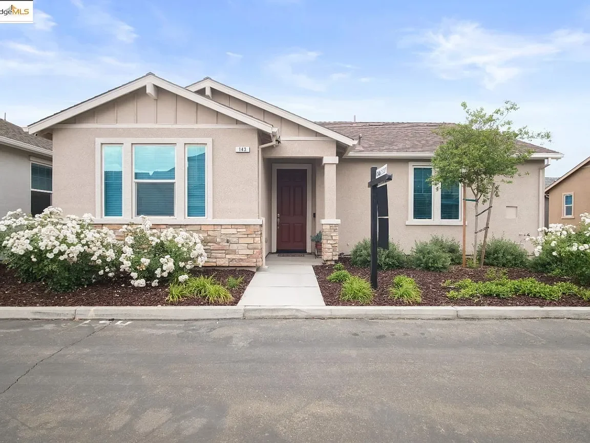 Front view of a modern single-family house with a small garden, concrete walkway, and a purple front door. The house has beige walls, a sloped roof, and large windows with white frames. A black mailbox is near the sidewalk.