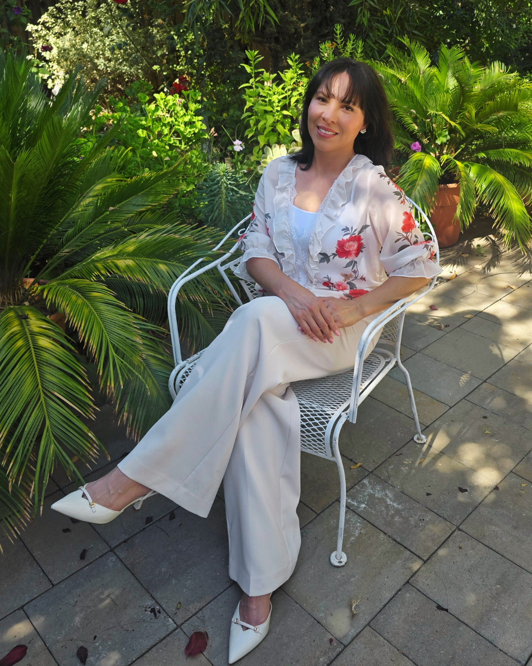 Woman sitting on white metal bench in a lush garden with green plants and potted flowers, wearing a floral blouse and white trousers, smiling at the camera.