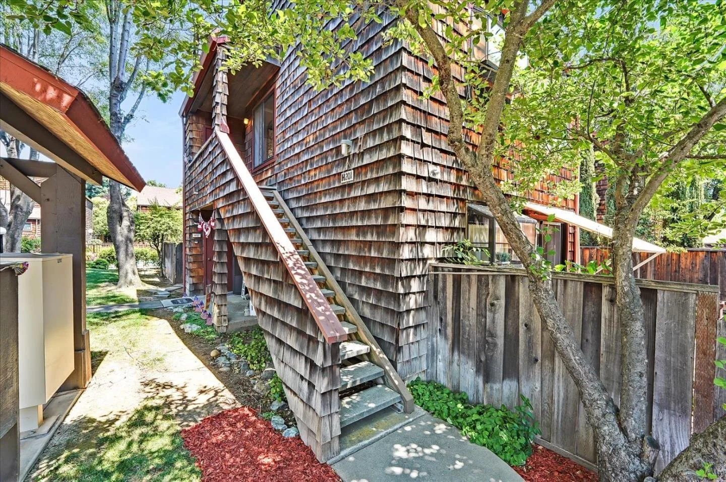 The side of a wooden house with shingle siding, a staircase leading to the second floor, and a backyard with trees and a wooden fence.