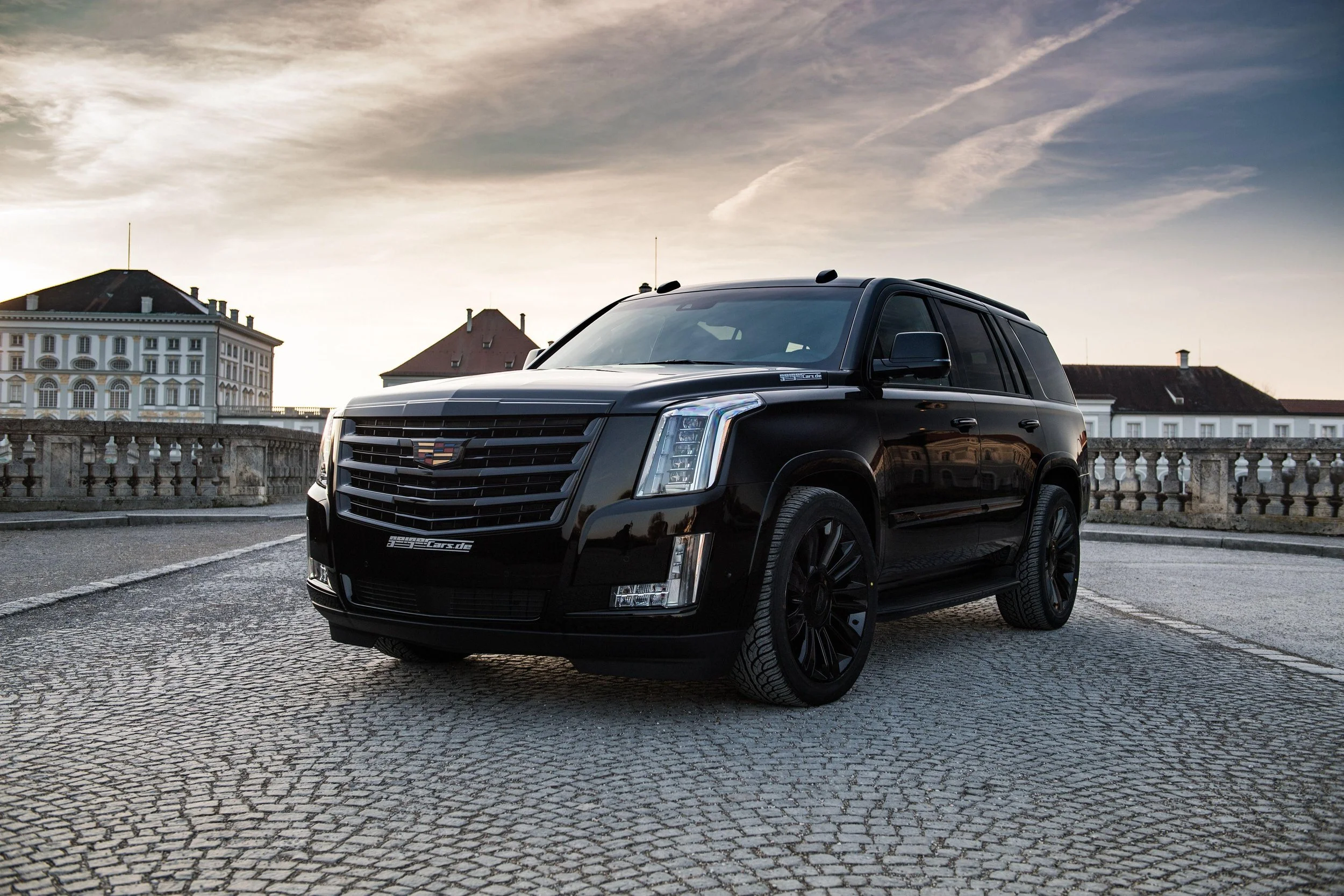A black Cadillac SUV parked on a cobblestone street with historic buildings and a cloudy sky in the background.