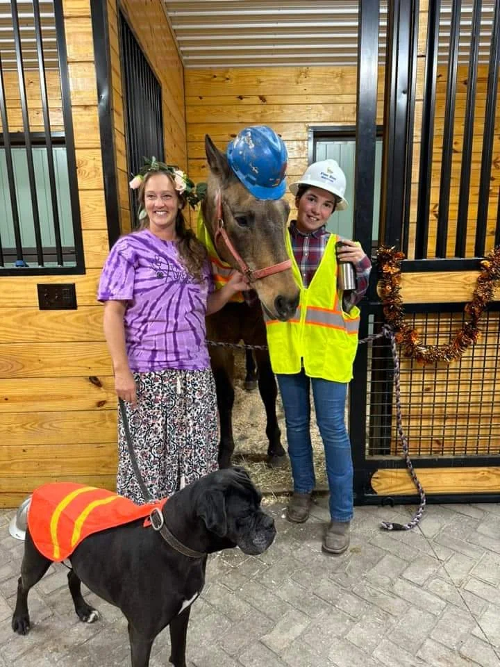 Two girls, one wearing a purple tie-dye shirt and floral skirt, and the other in a plaid shirt with a white safety helmet and yellow safety vest, stand inside a barn with a horse between them, all smiling. There is also a black dog wearing an orange safety vest standing in front. The barn has wooden walls and metal bars, with decorations including a wreath.