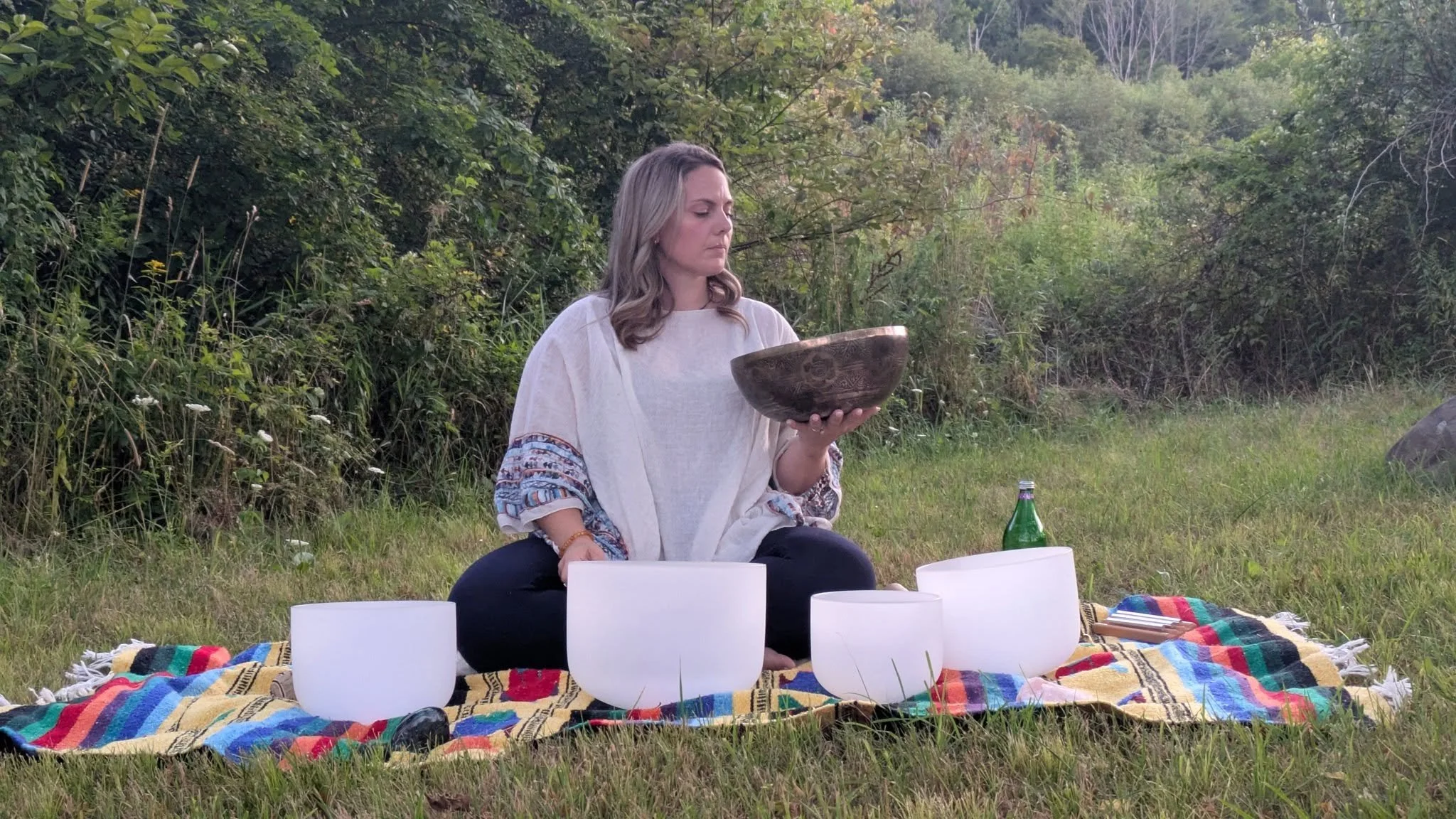 A woman sitting cross-legged on a colorful woven blanket outdoors, holding a singing bowl and surrounded by other similar bowls.. The background shows a lush, green wooded area  at NorthWoods Farm, Dushore, PA
