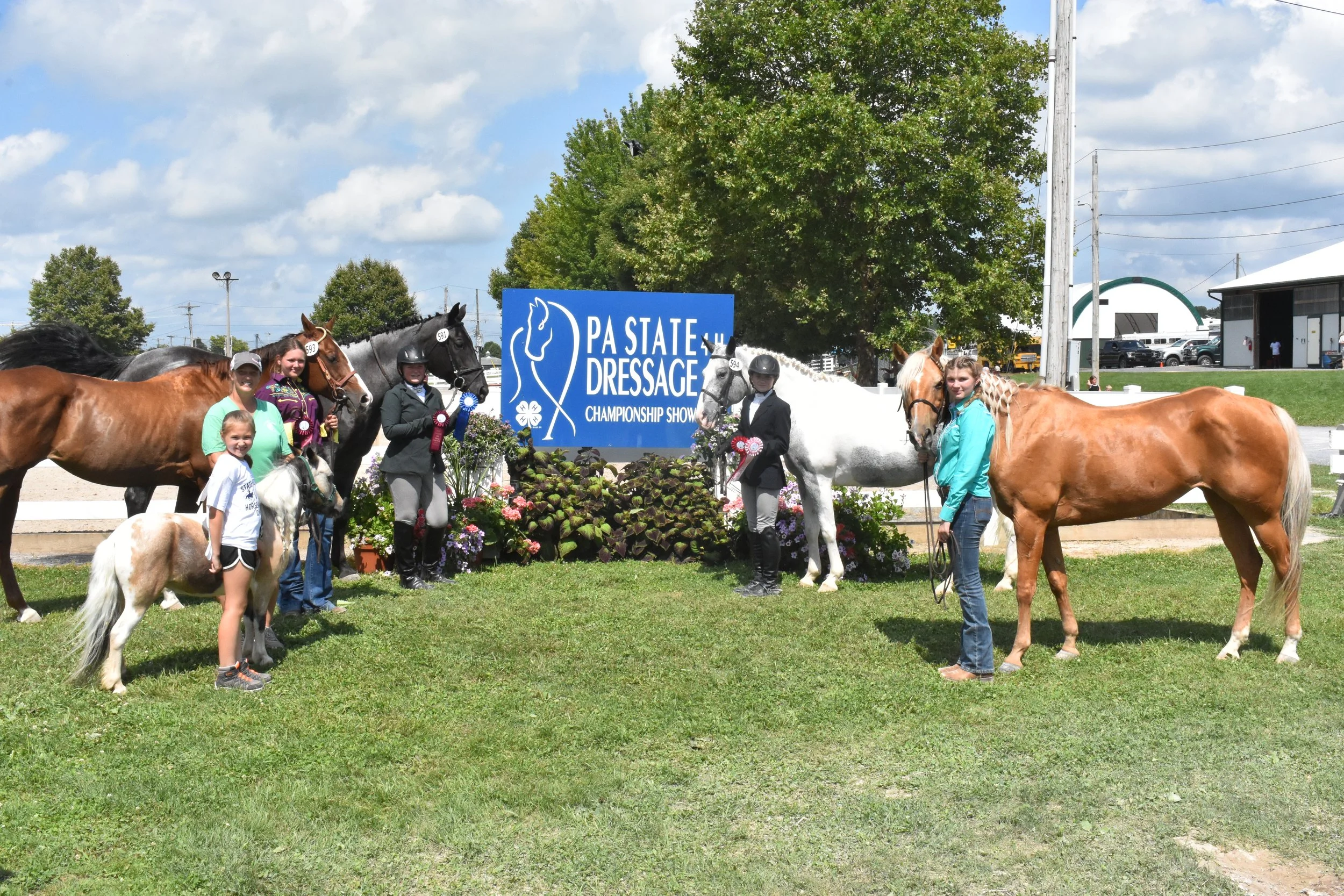 People at a horse show, some with ribbons, standing with their horses in front of a sign that reads 'PA State Dressage Championship Show', with trees, buildings, and vehicles in the background.