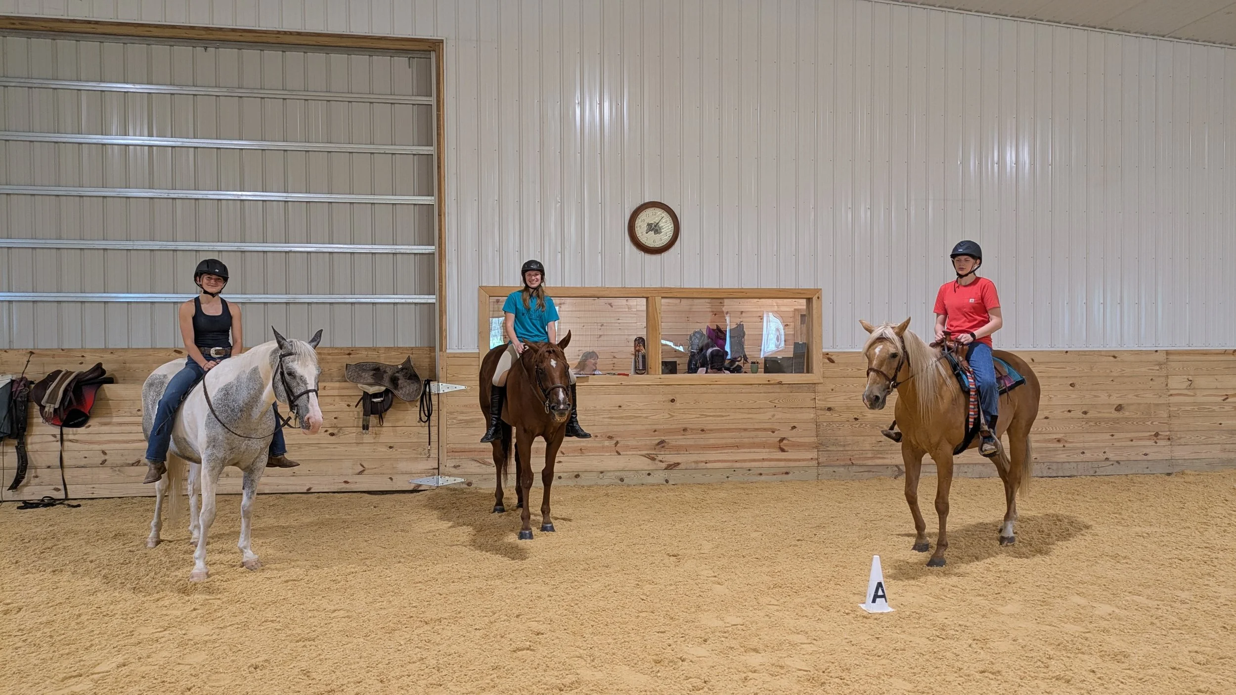 Three young women wearing horse riding helmets sitting on horses in an indoor riding arena with a wooden fence and a clock on the wall. Lessons or clinic.  at NorthWoods Farm, Dushore, PA