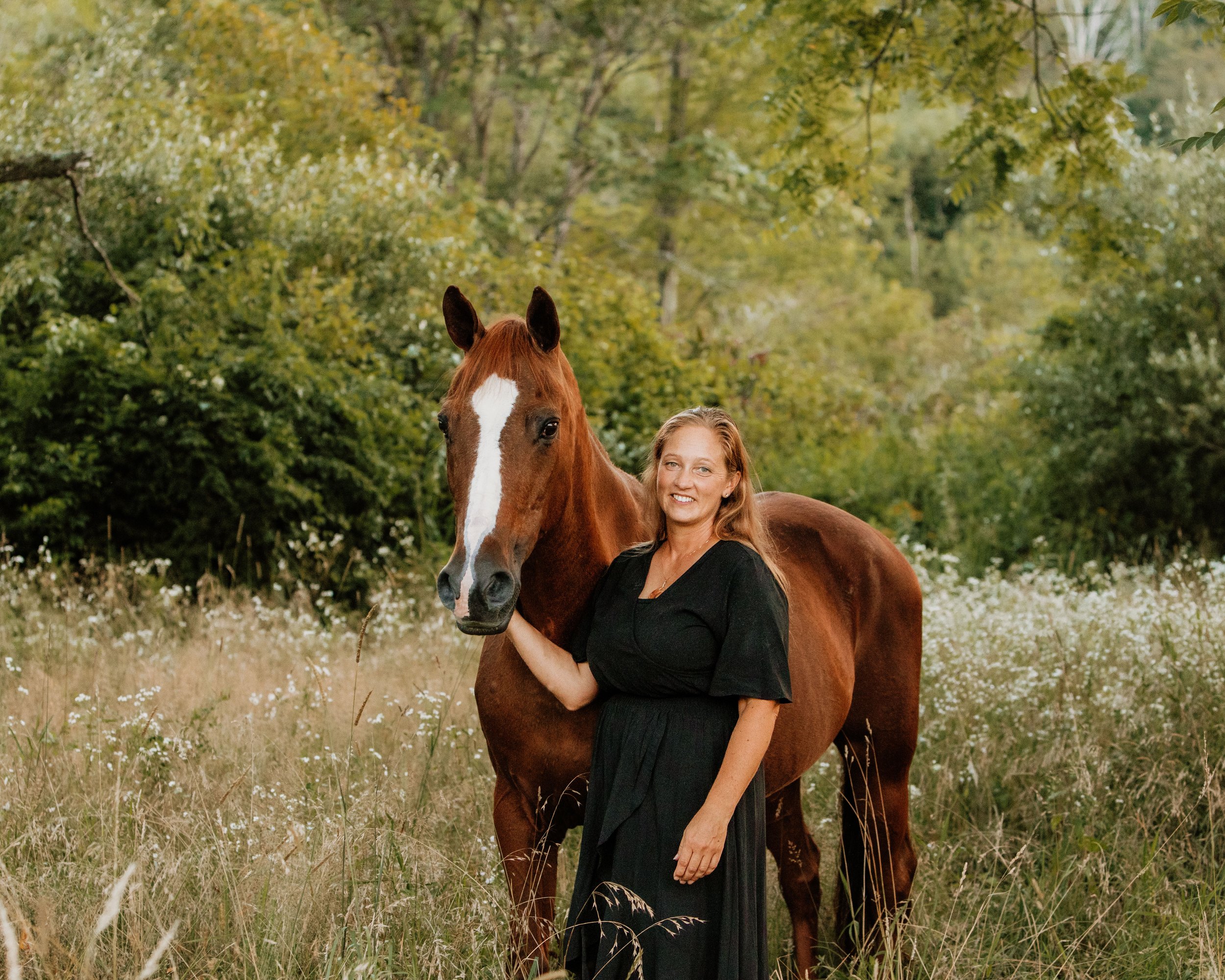 A woman with long reddish hair standing next to a brown horse in a grassy field with trees and greenery in the background.
