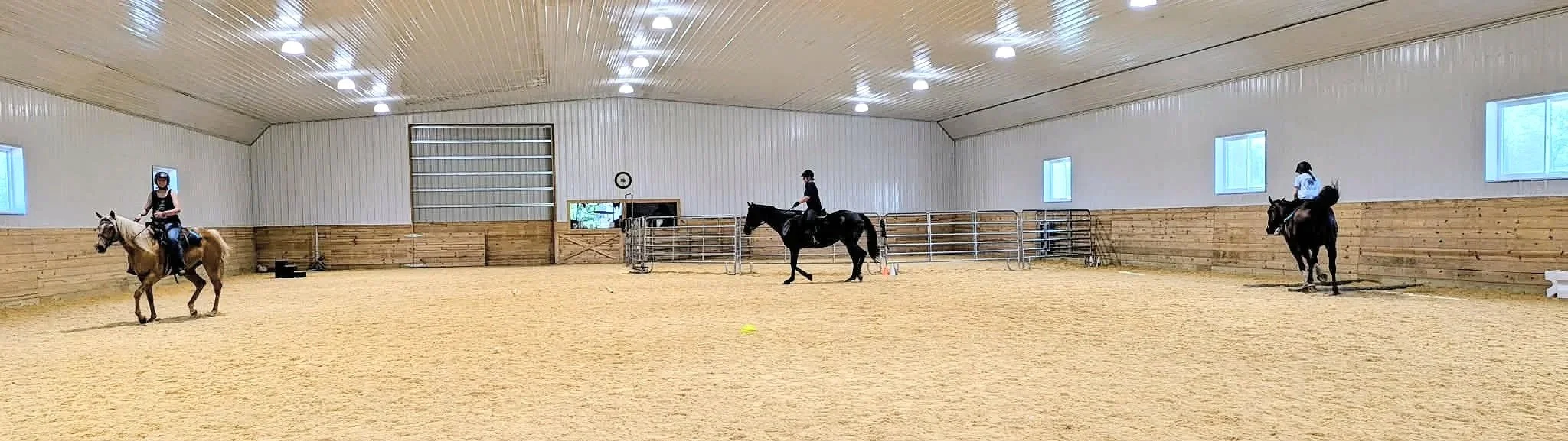 Three horseback riders in an indoor riding arena with wooden walls, a sandy floor, and windows on the walls. Small group lesson with games on horseback.