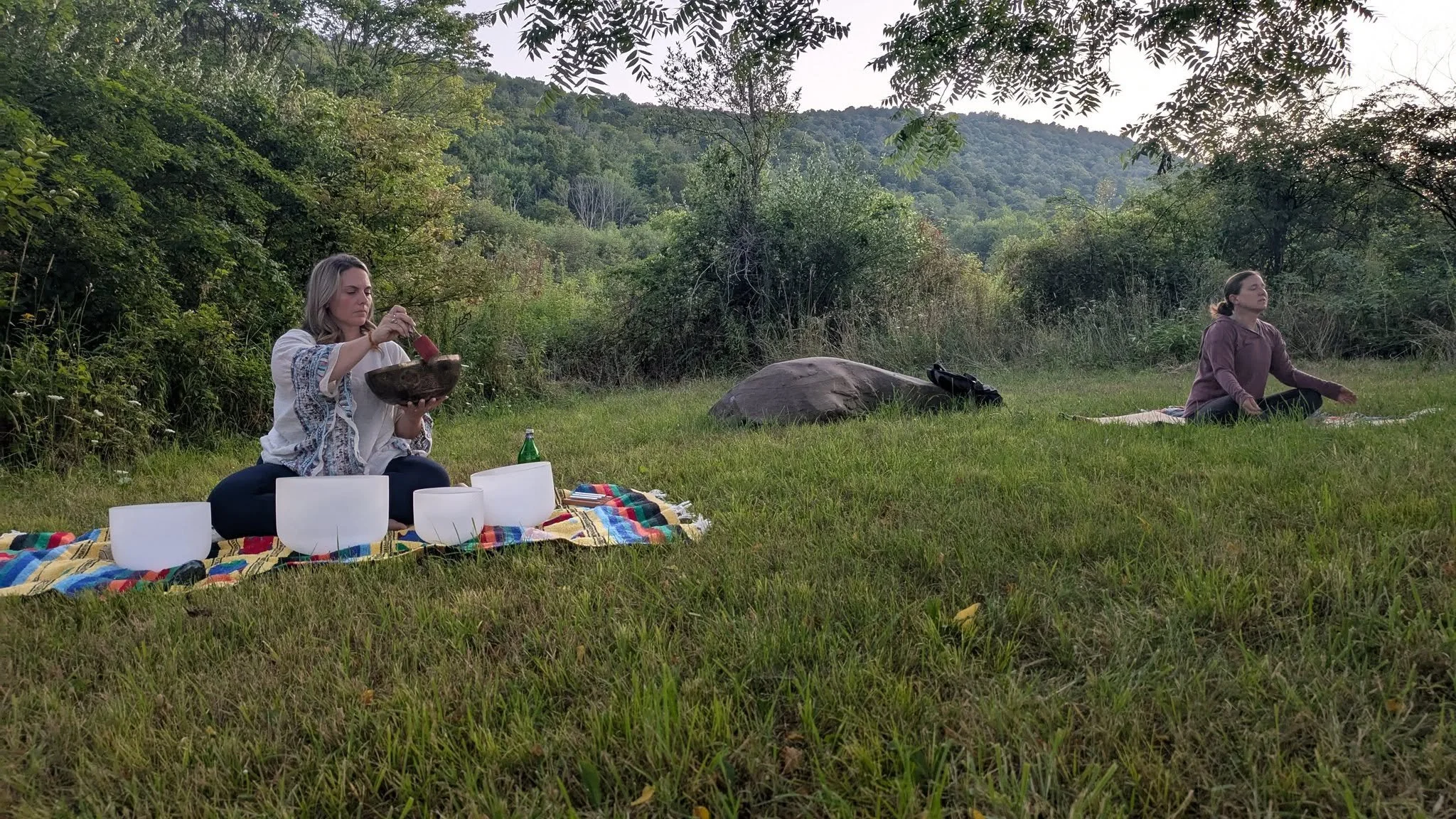 Two women practicing yoga outdoors in a forest clearing, one sitting on a colorful blanket with singing bowls, the other seated on a yoga mat with eyes closed, surrounded by trees and mountains at NorthWoods Farm, Dushore, PA