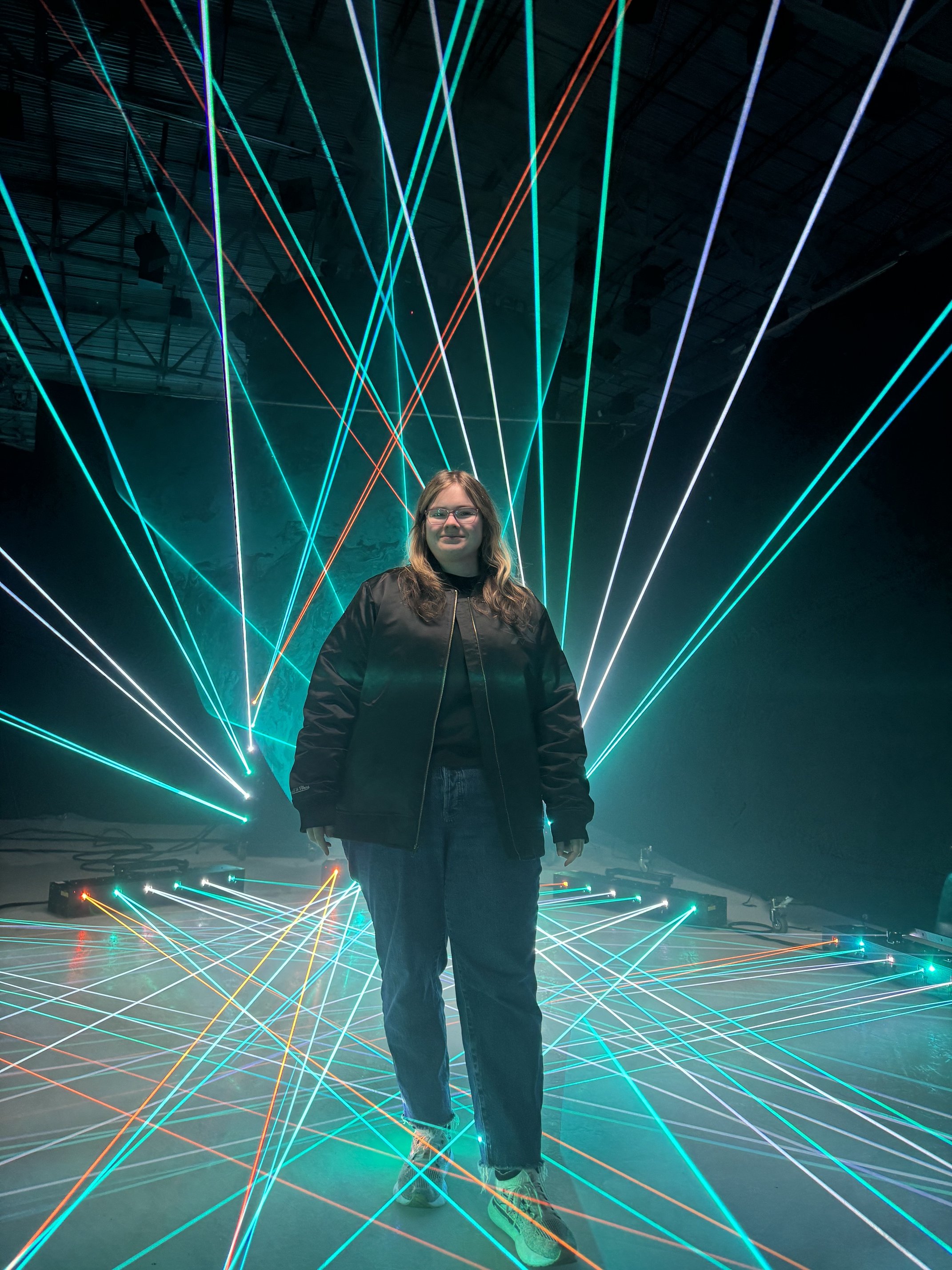 A woman with glasses and brown hair, wearing a black jacket and jeans, stands in front of a background of colorful laser lights on the floor and ceiling in a dark, indoor space.