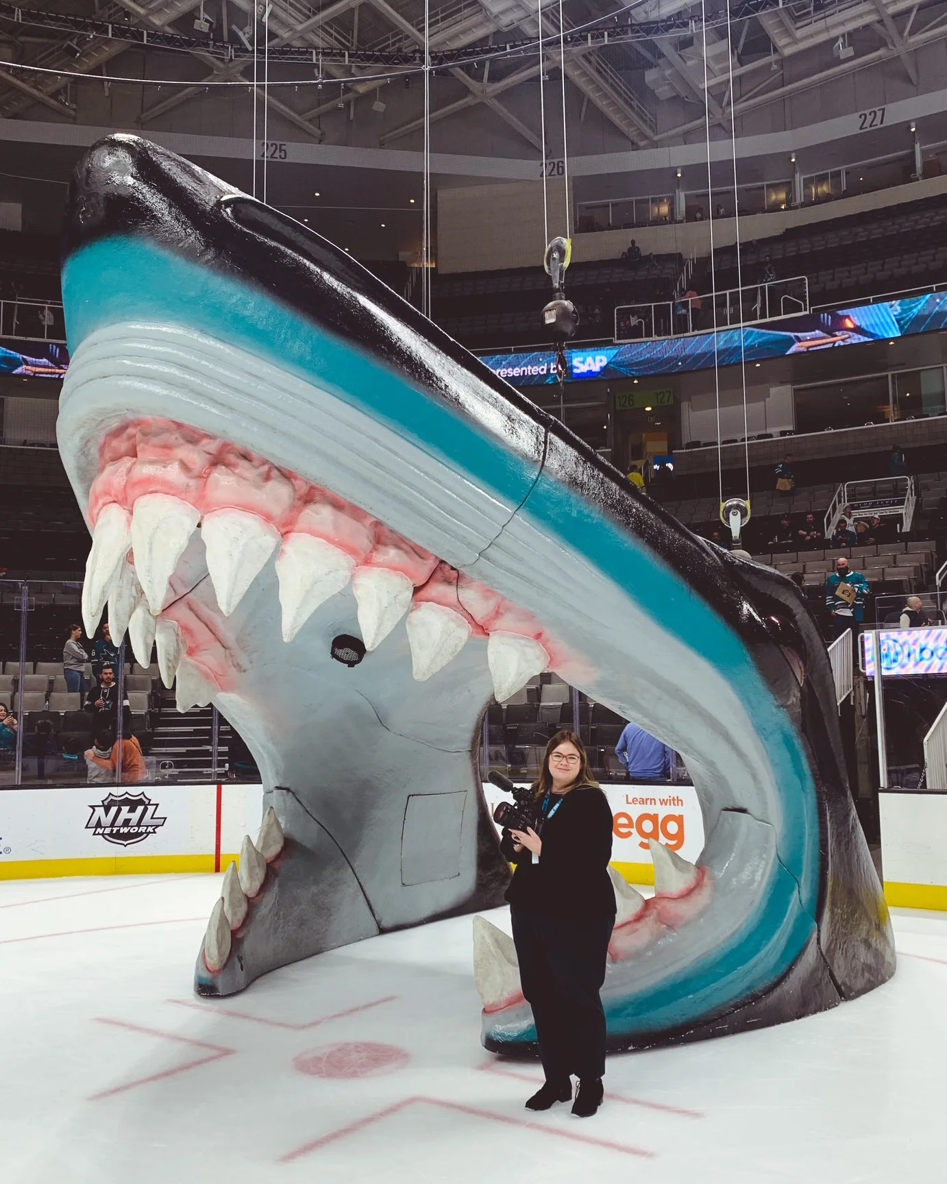 A woman standing inside the open mouth of a large shark sculpture on an ice rink. The shark has a black, white, and blue body, with sharp teeth and an intimidating appearance. The surrounding area appears to be an indoor arena with spectators and advertising banners.