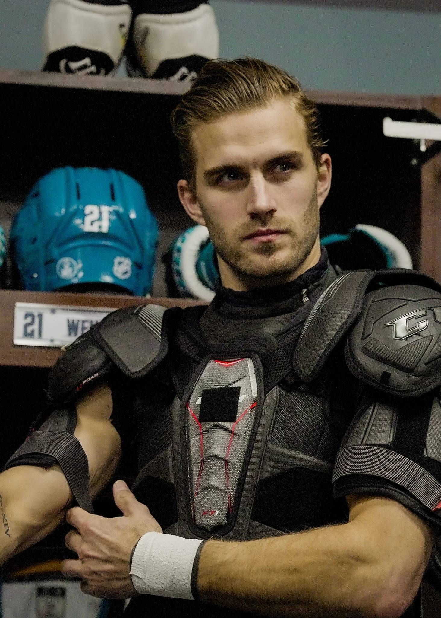 A man with slicked back hair and a serious expression wearing a black and gray football shoulder pad, white wristband, and a black shirt, standing in a locker room. Behind him on the shelf are white and black shoes and a blue football helmet with a w