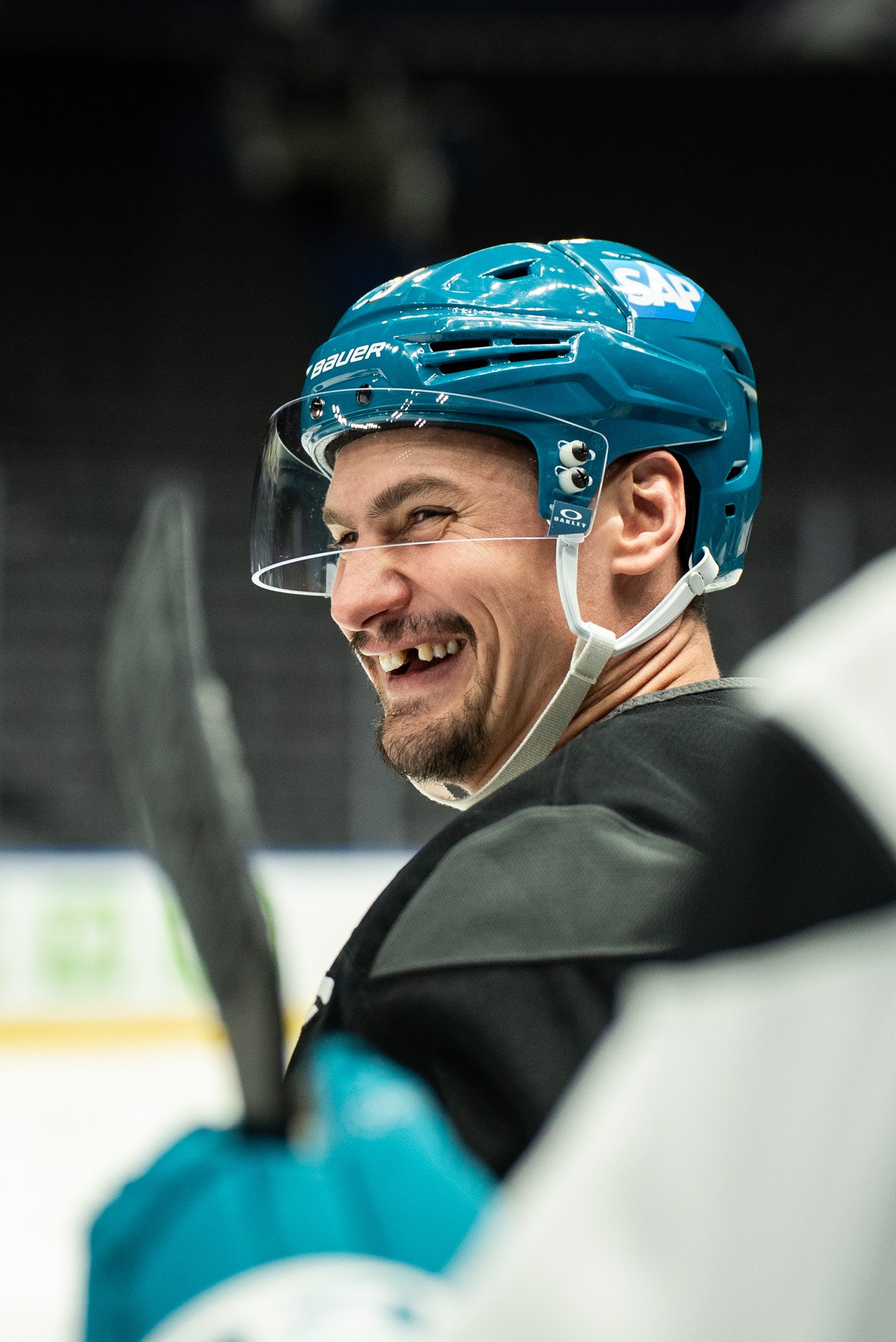 A male ice hockey player with a beard, wearing a blue helmet with a white logo, a clear face shield, and black and teal gear, smiling while looking to the side.