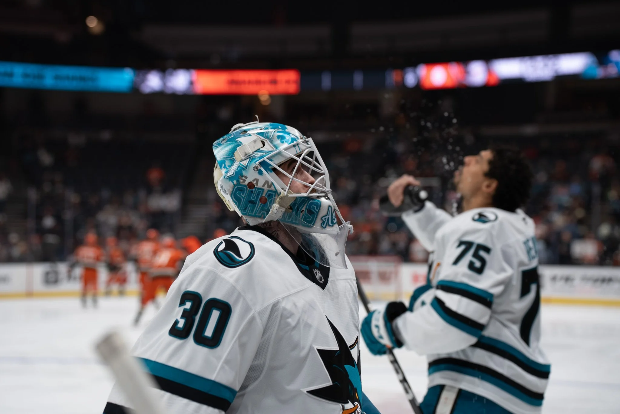 Ice hockey players on the ice during a game, with one player in focus wearing a white jersey with teal and black accents, and a goalie wearing a matching helmet and jersey, in an arena with blurred spectators and scoreboard.
