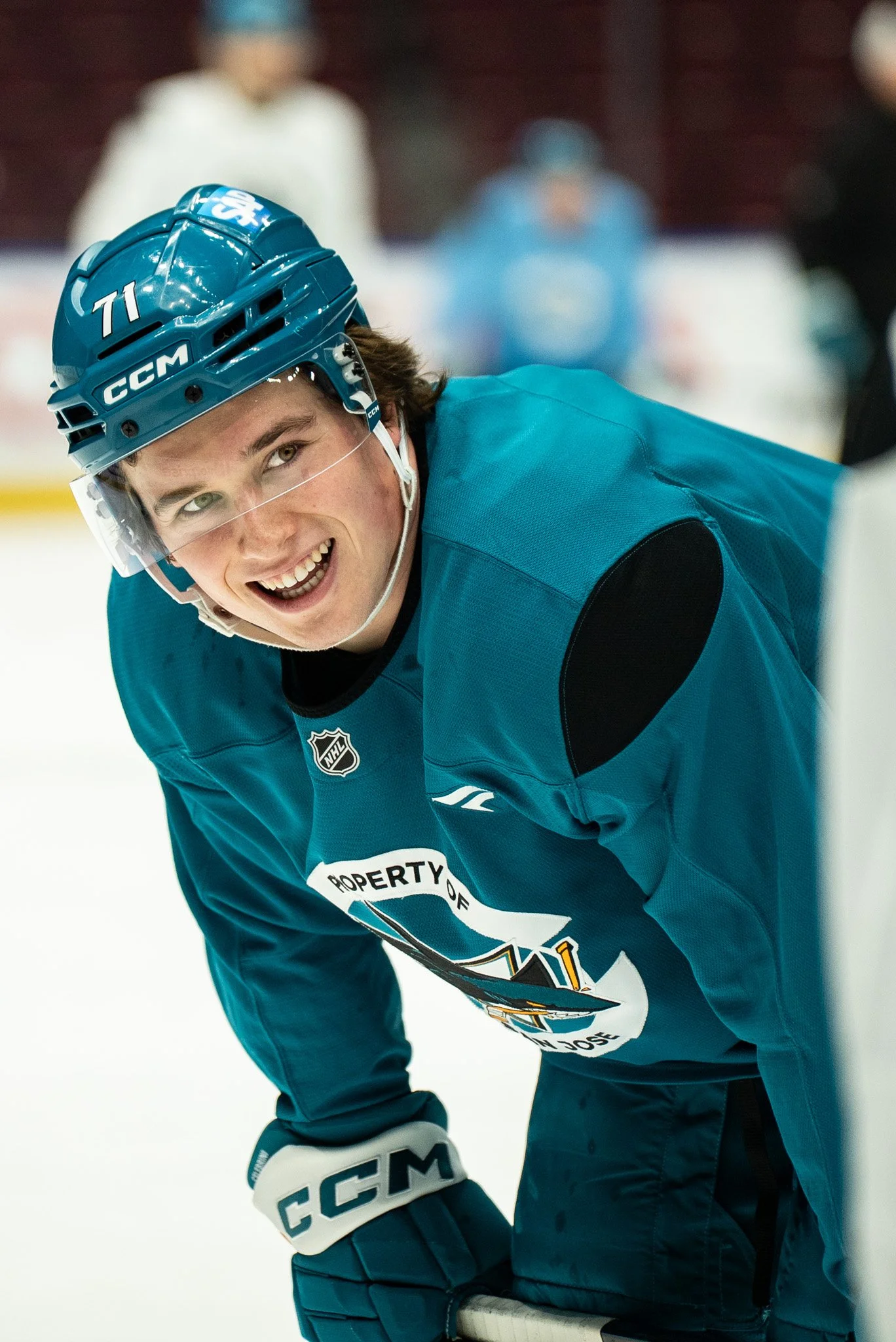 Young male hockey player smiling on ice, wearing a blue jersey, helmet, and gloves, with a partially visible hockey stick.