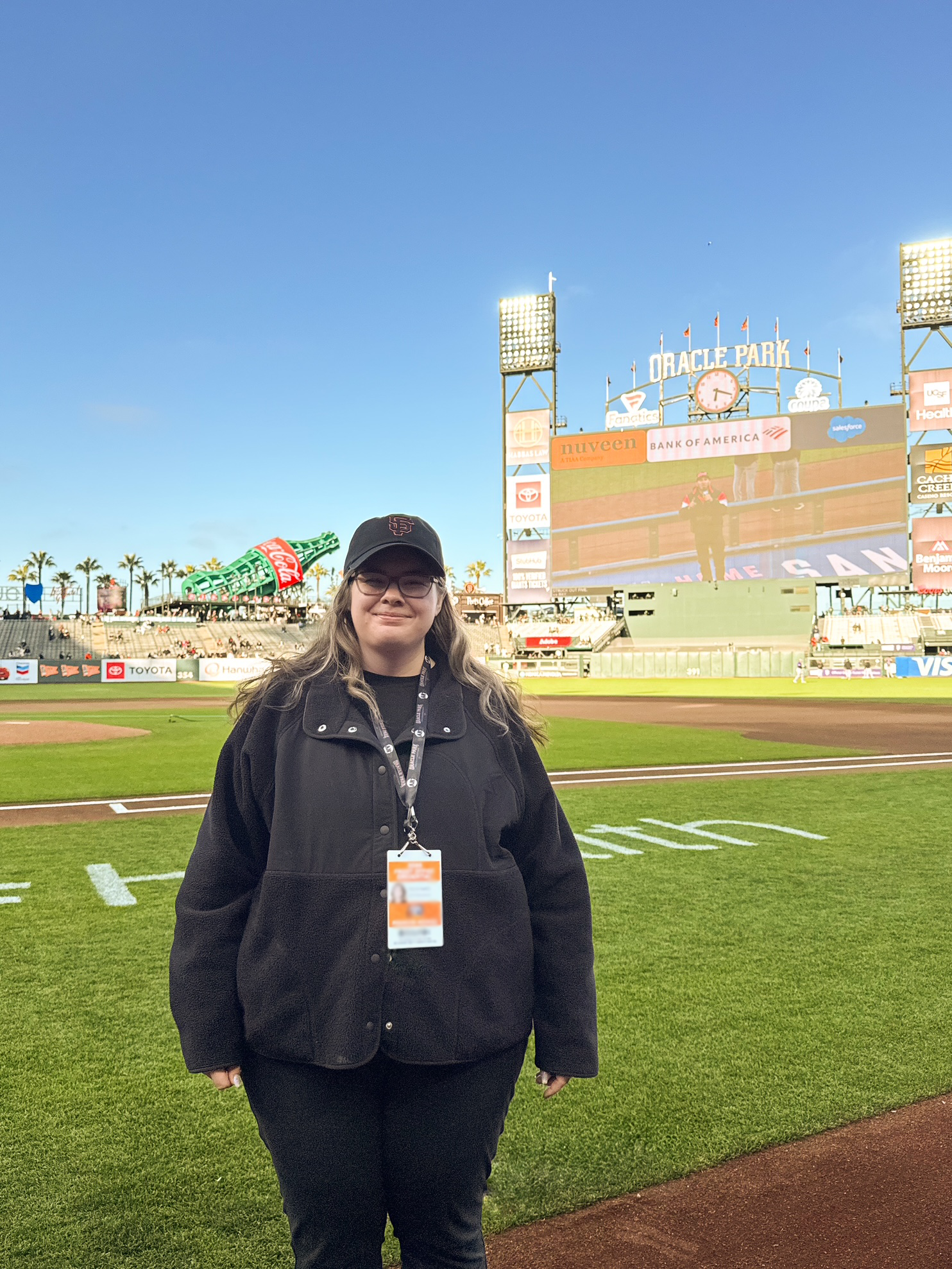 A woman standing on a baseball field in front of a large scoreboard and a giant Coca-Cola bottle sculpture at Oracle Park during the day.