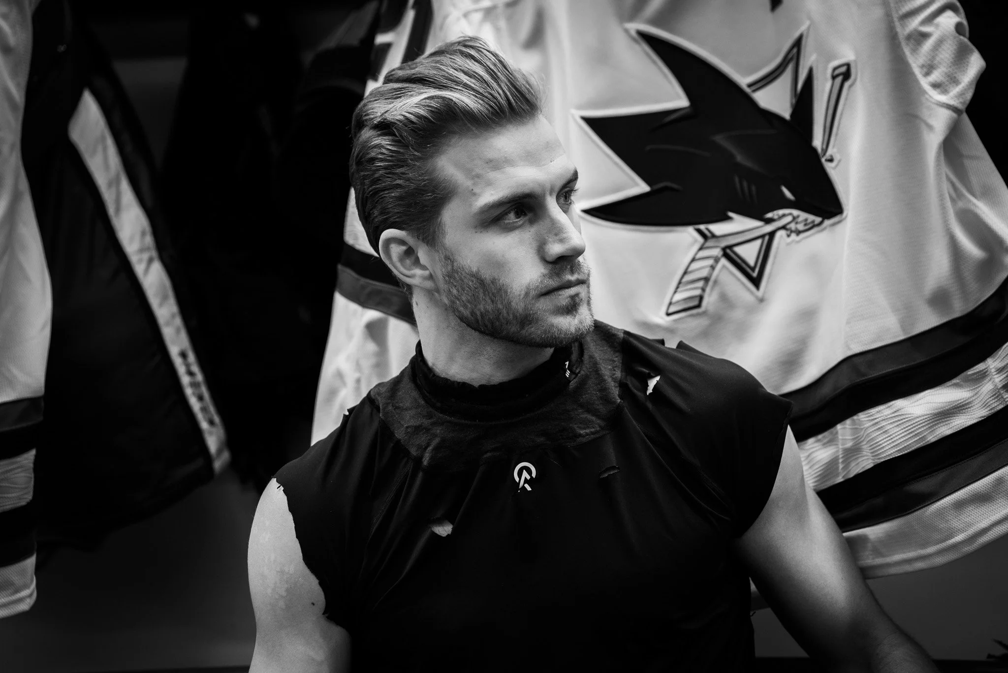 A young man with styled hair and a beard sitting with hockey players and a hockey jersey in the background.