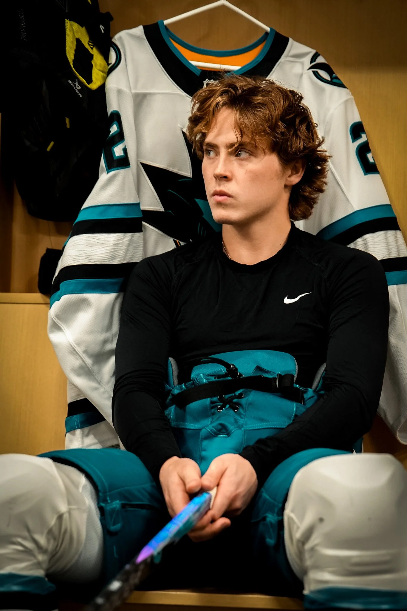 A young man with curly hair sitting in a locker room in front of a hockey jersey and holding a colorful hockey stick.