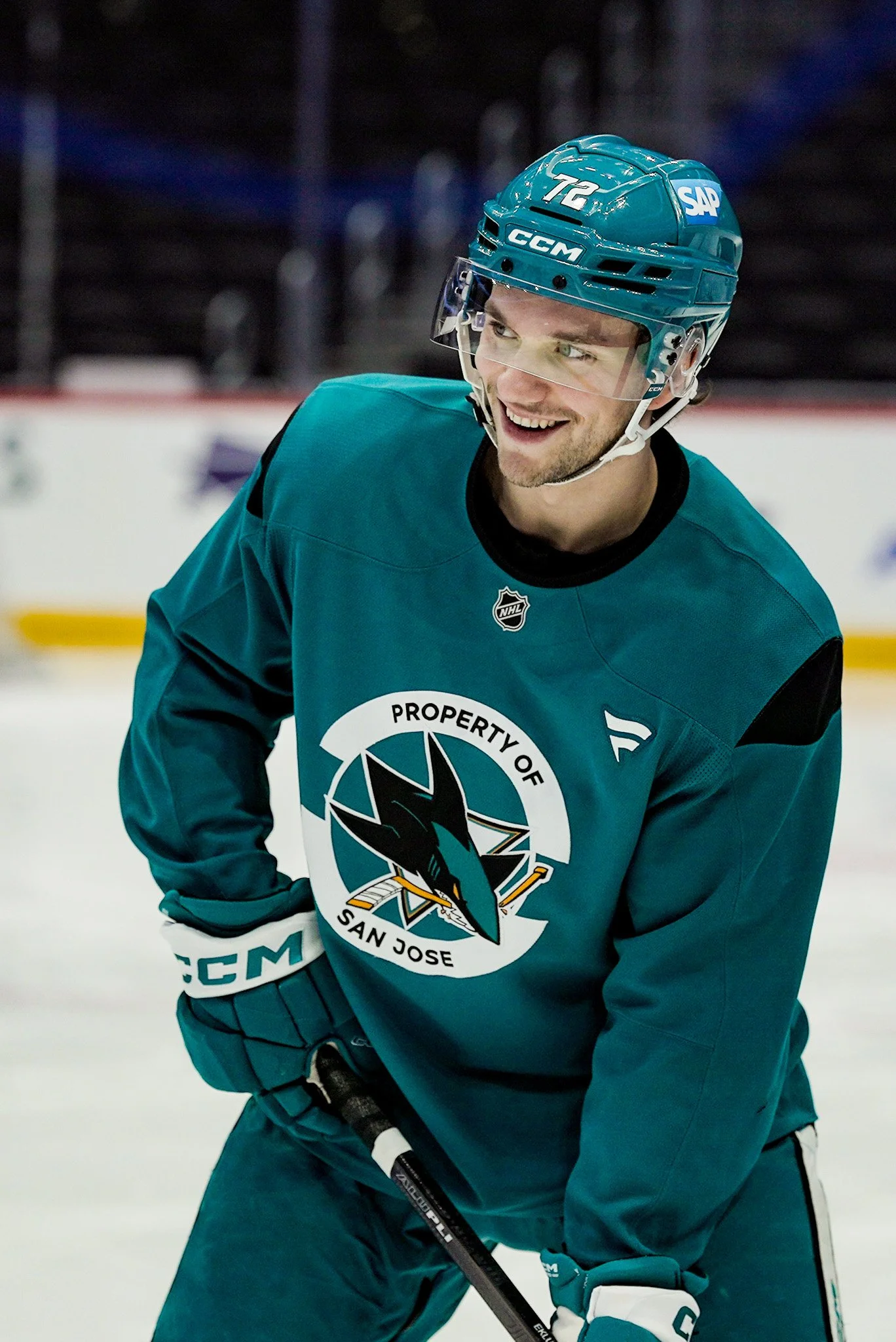 An ice hockey player in teal jersey smiling on the ice rink, wearing a blue helmet with the number 72 and stickers, holding a hockey stick, with the logo of San Jose Sharks on his jersey.