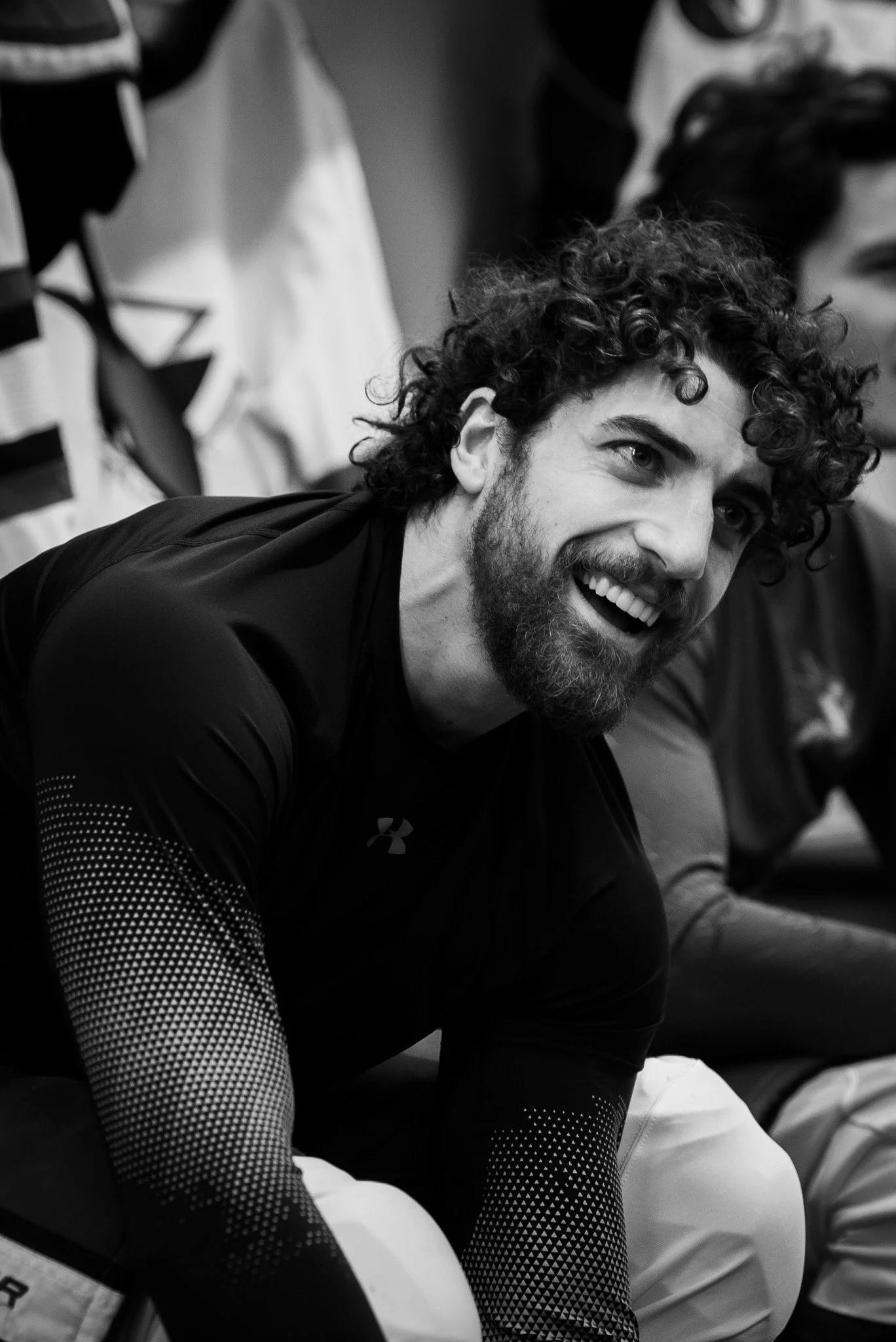 A man with curly hair and a beard smiling while sitting on the floor, wearing a black Under Armour shirt with patterned sleeves. Mario Ferraro, San Jose Sharks