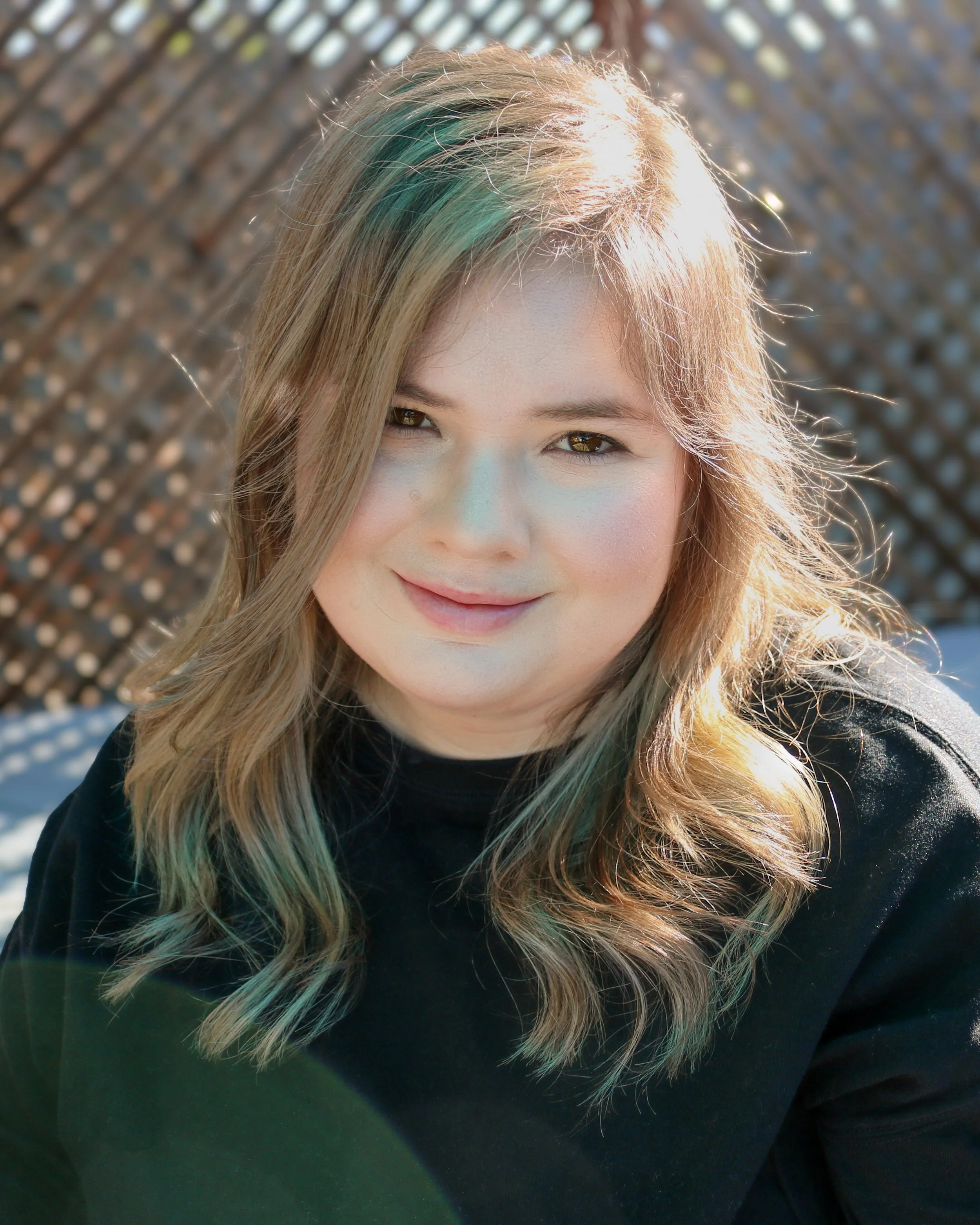 A woman with long blonde hair with green streaks, smiling, wearing a black top, with a background of a metallic fence and outdoor setting.