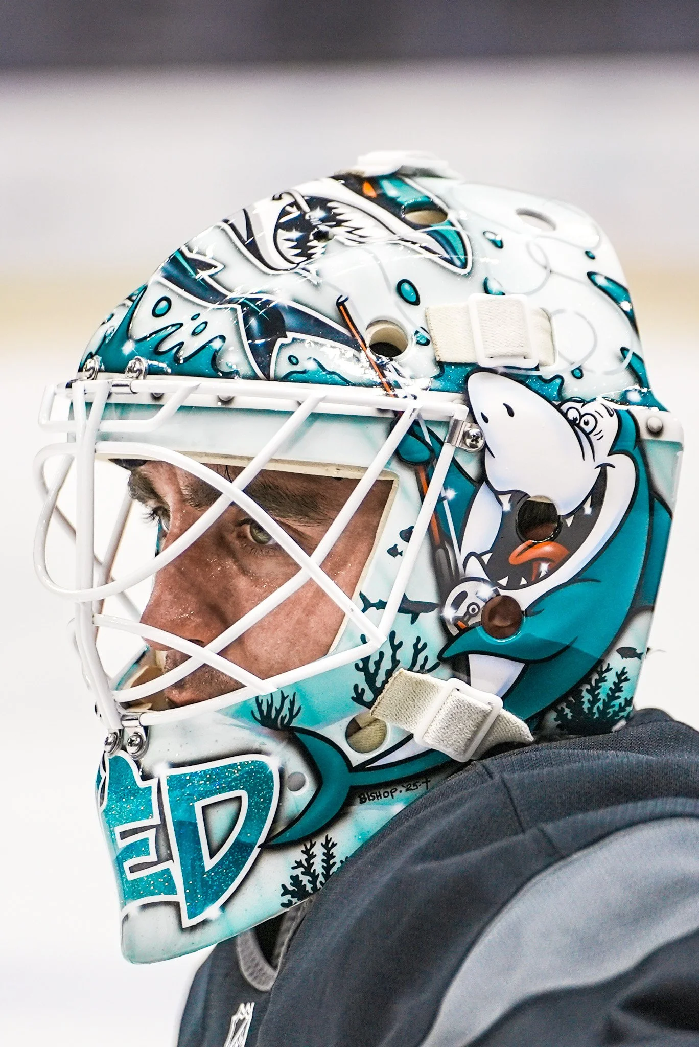 A hockey goalie wearing a helmet with a cartoon shark design, partially visible face, and ocean-themed graphics.