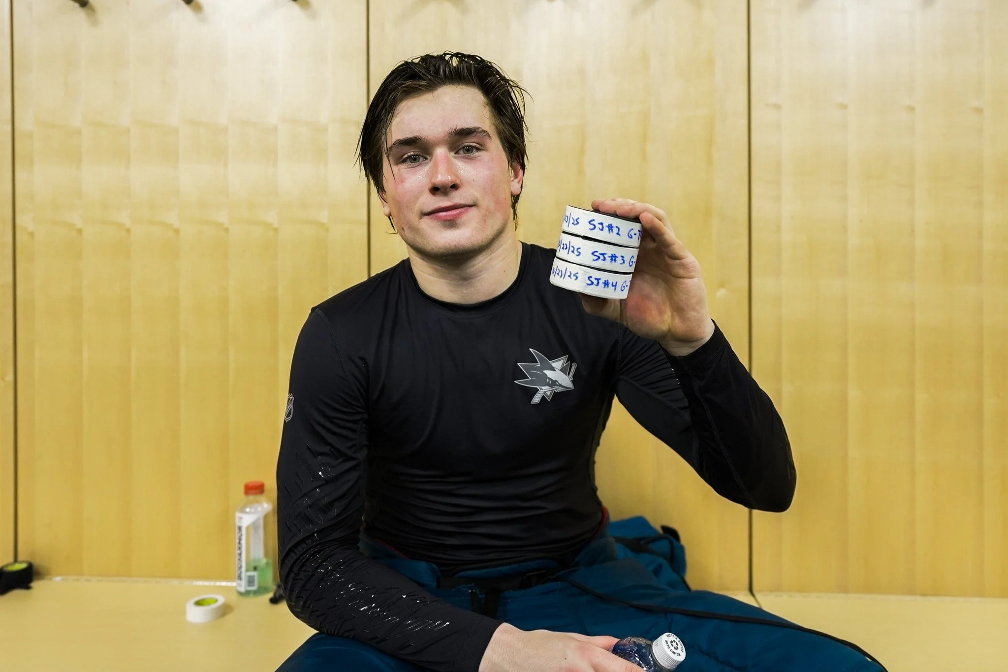A young man with dark hair, wearing a black sports jersey, sitting and smiling while holding a rolled piece of tape with writing on it. The background is a plain yellowish wall with vertical panels, with sports bottles and tape visible on the table n