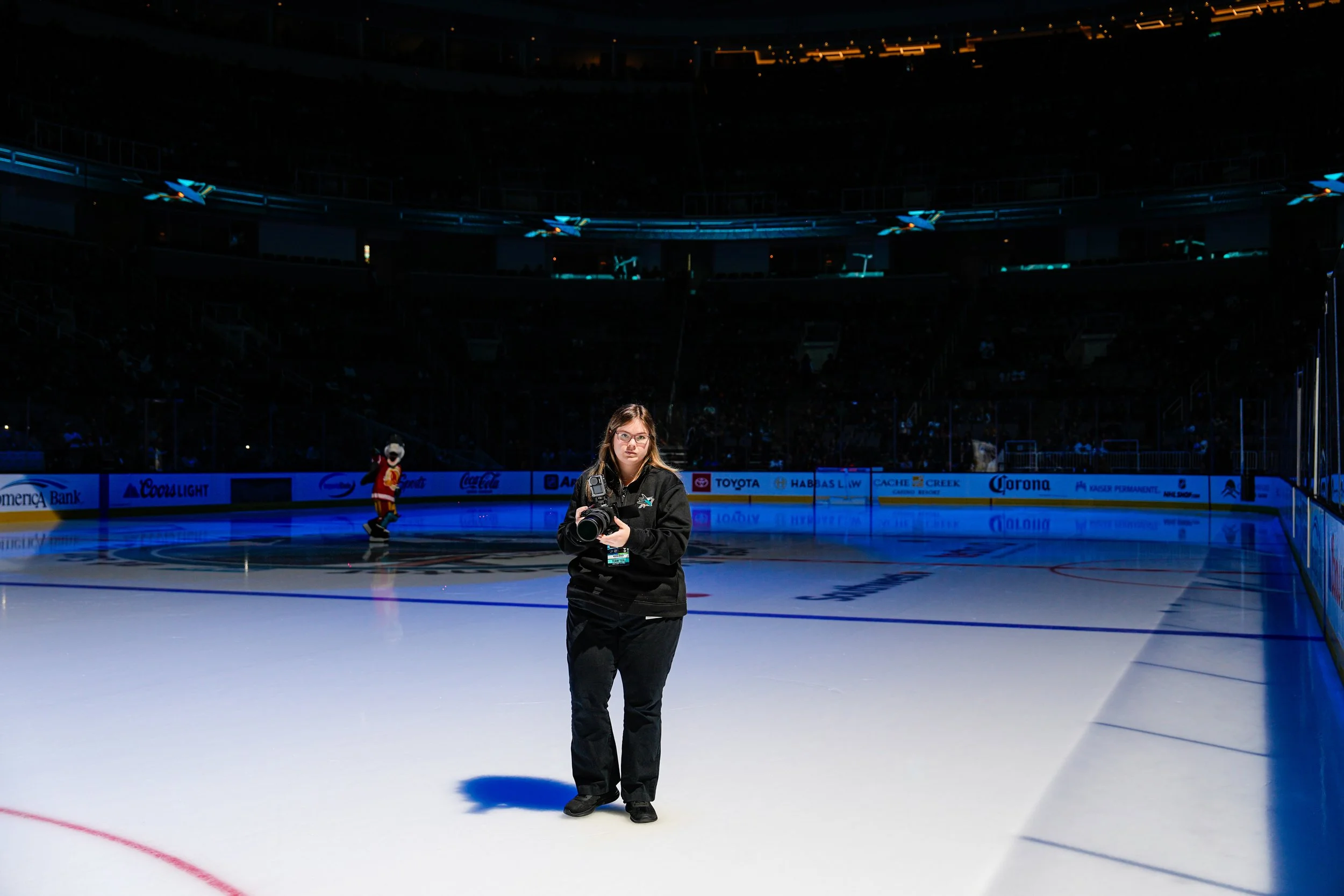 An indoor ice hockey rink with a young woman standing on the ice holding a camera. There is a mascot in a hockey jersey in the background. The rink is surrounded by a barrier with advertisements, and the seating area is dark with only a few lights visible.