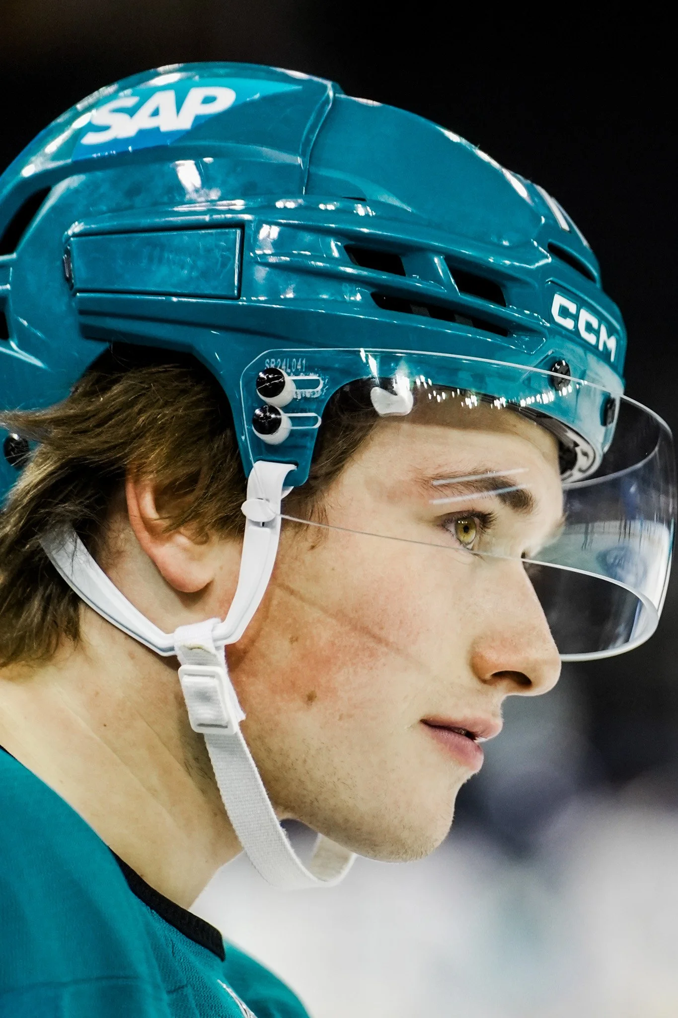 Close-up of an ice hockey player wearing a blue helmet with SAP and CCM logos, clear visor, and teal jersey. Macklin Celebrini, San Jose Sharks