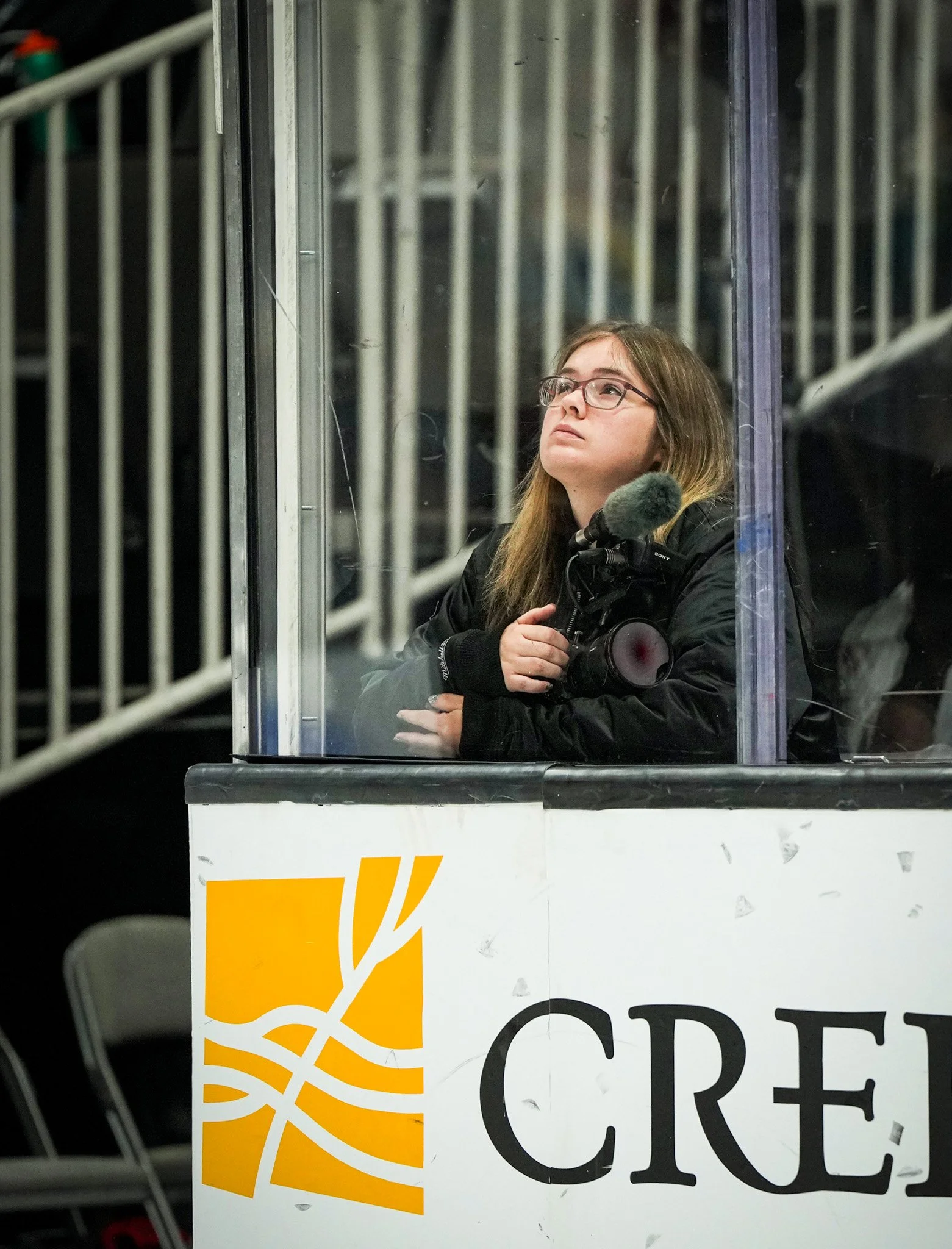 A young woman with brown hair and glasses, holding a camera, sitting behind a barrier with a sign that has a yellow logo and the partially visible word "CRES" in black text.