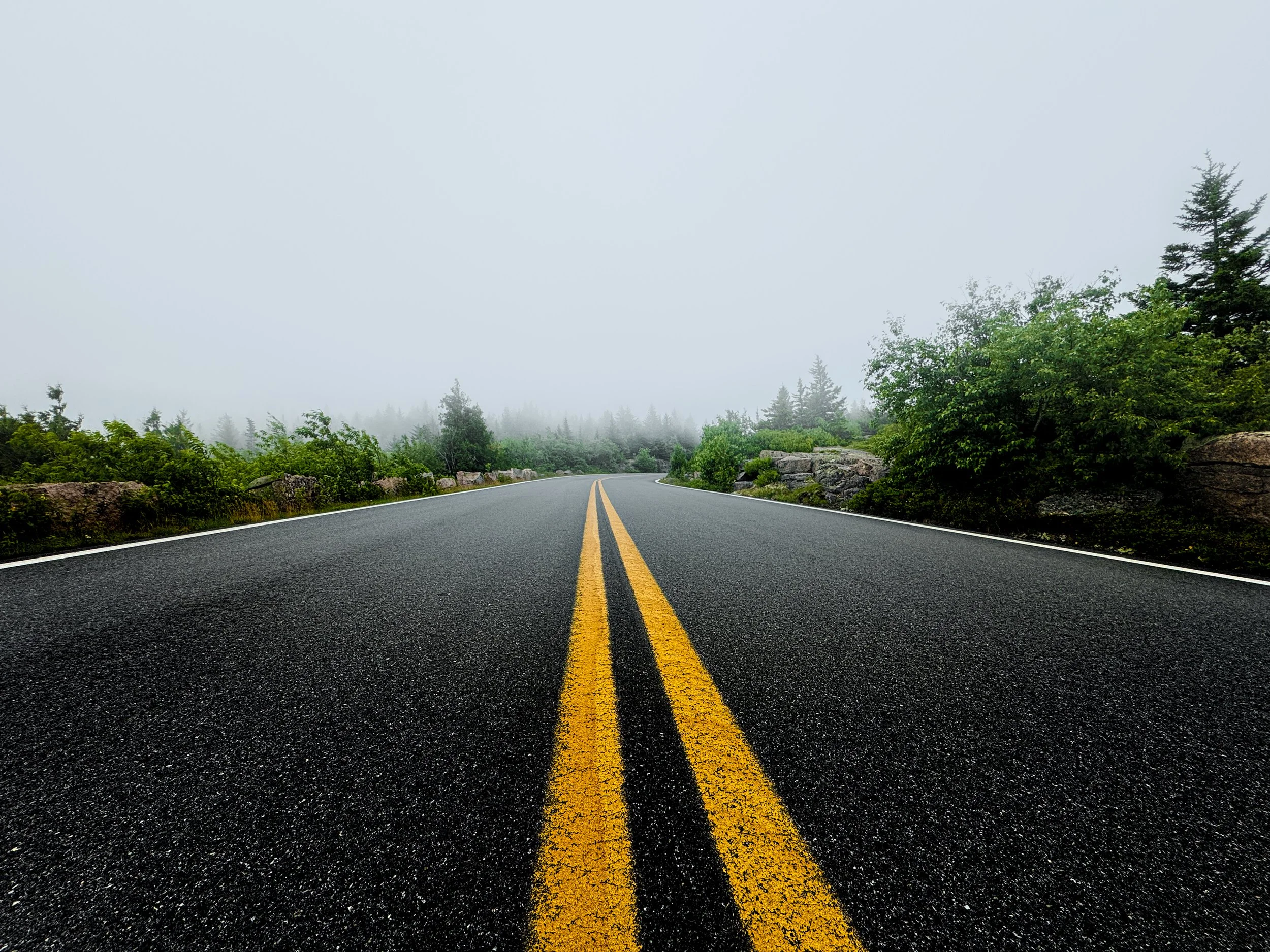 A two-lane road with yellow double lines running through a foggy forest landscape.