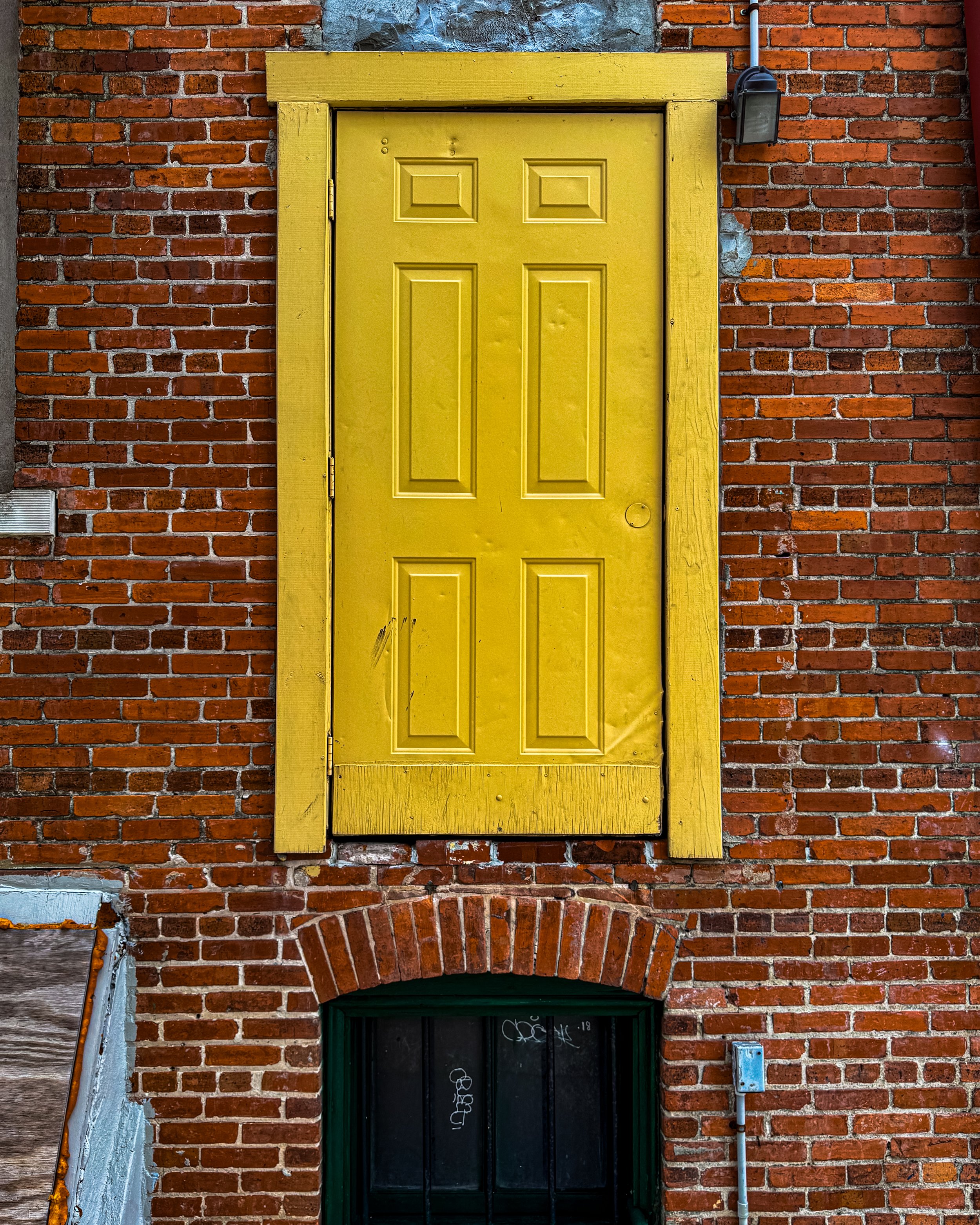 A yellow wooden door set in a red brick wall, with black window bars and a small window at the top, framed by a matching yellow trim. The brickwork around the door shows signs of wear and weathering.