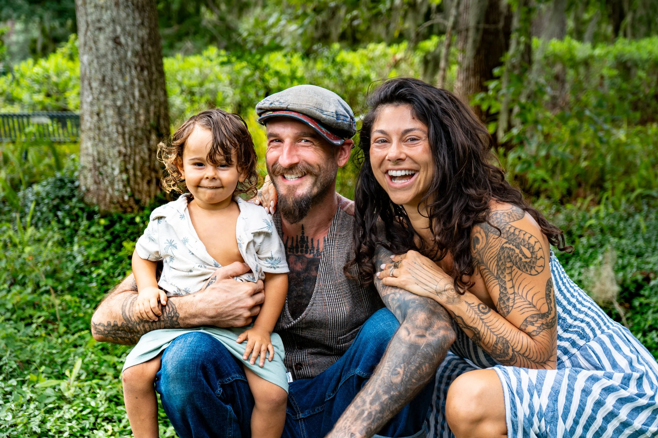 A group of three people, including a young child and two adults, sitting outdoors in a lush green forest or park, smiling and enjoying the moment.