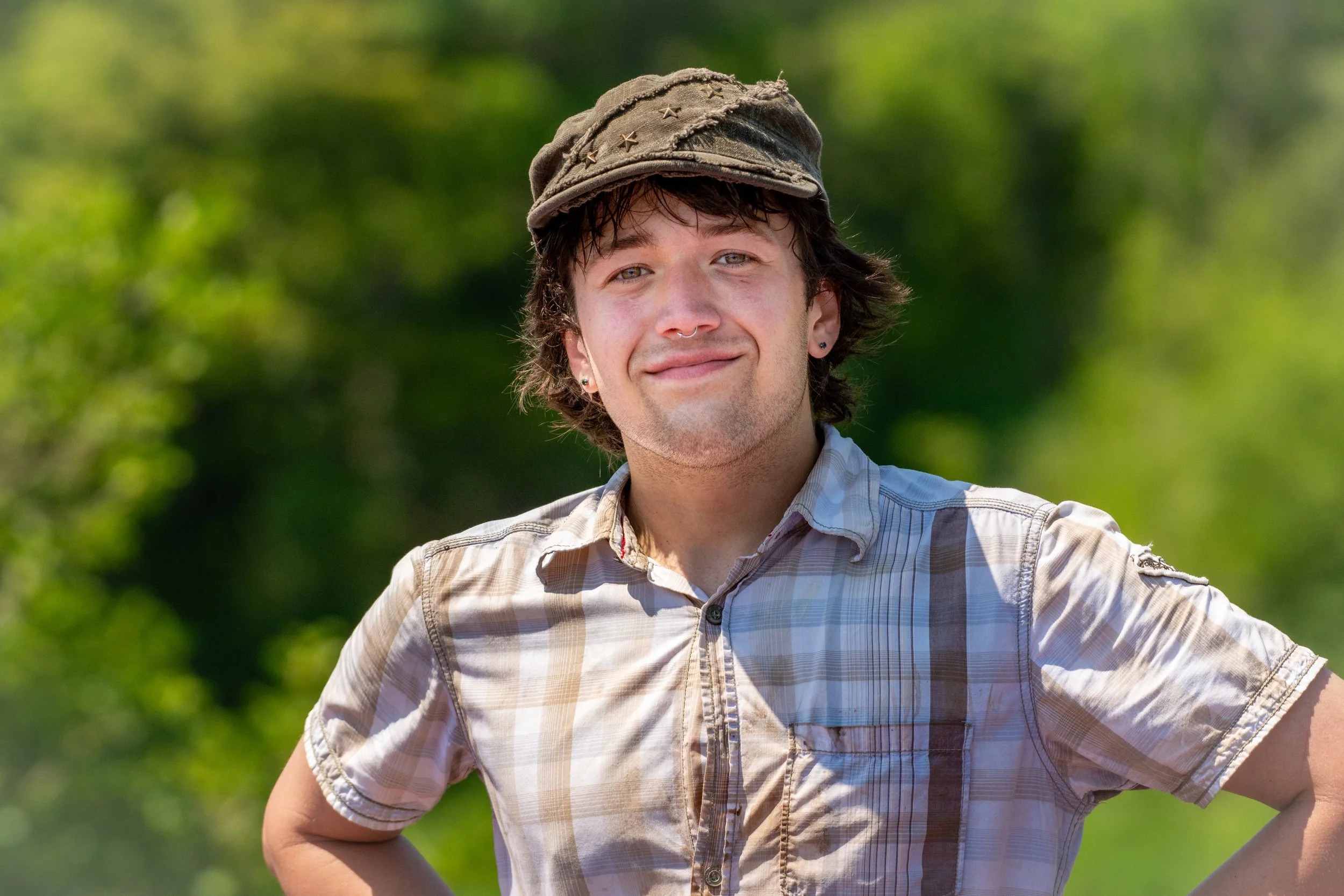 A young man with curly brown hair, earrings, and a slight smile, wearing a small weathered brown cap with star-shaped embellishments and a light-colored plaid short-sleeve shirt, standing outdoors with a blurred green background.