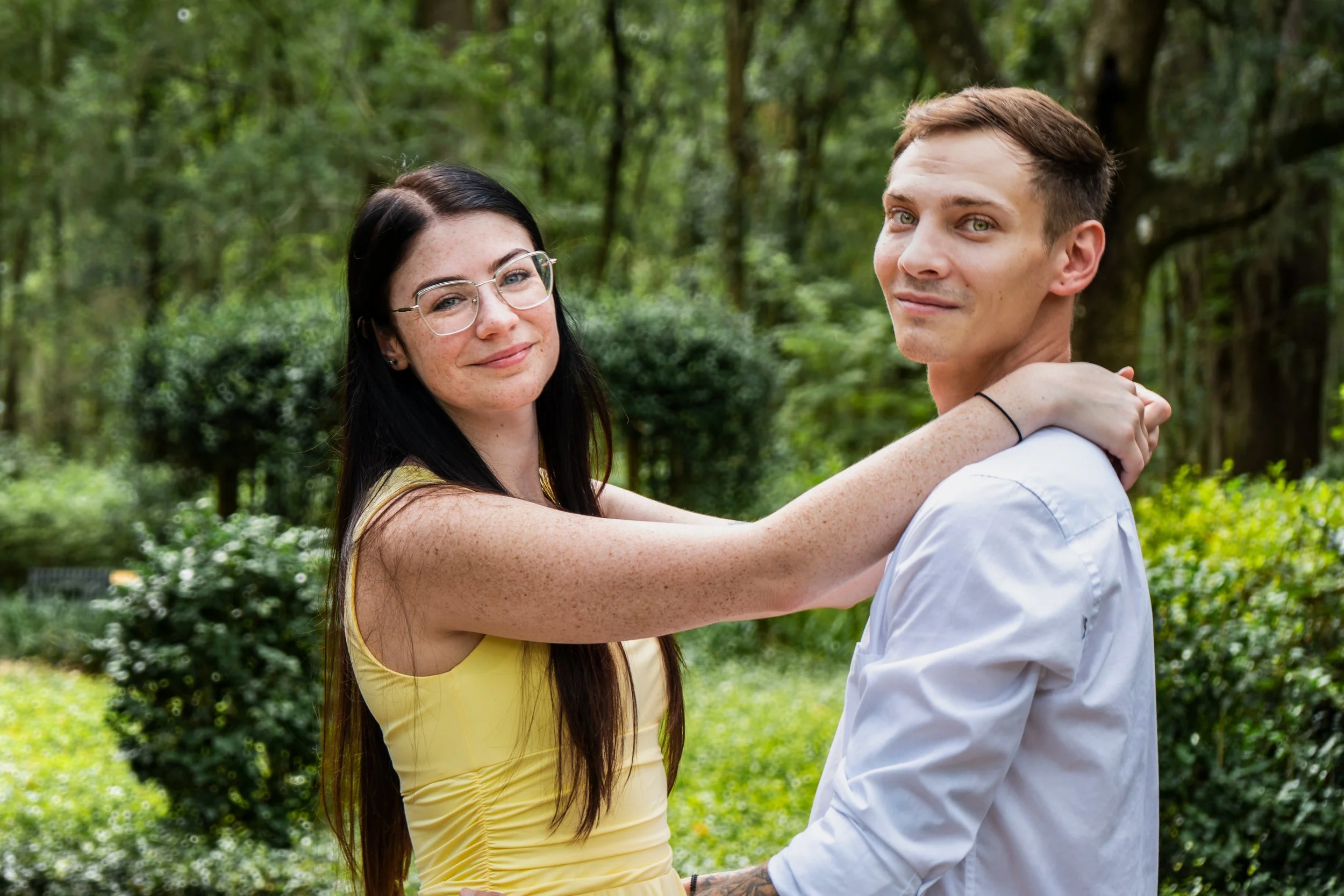 A young woman with long dark hair, glasses, and freckles wearing a yellow sleeveless top, holding a young man in a white shirt with short brown hair and freckles, standing outdoors in a green park with trees and bushes.
