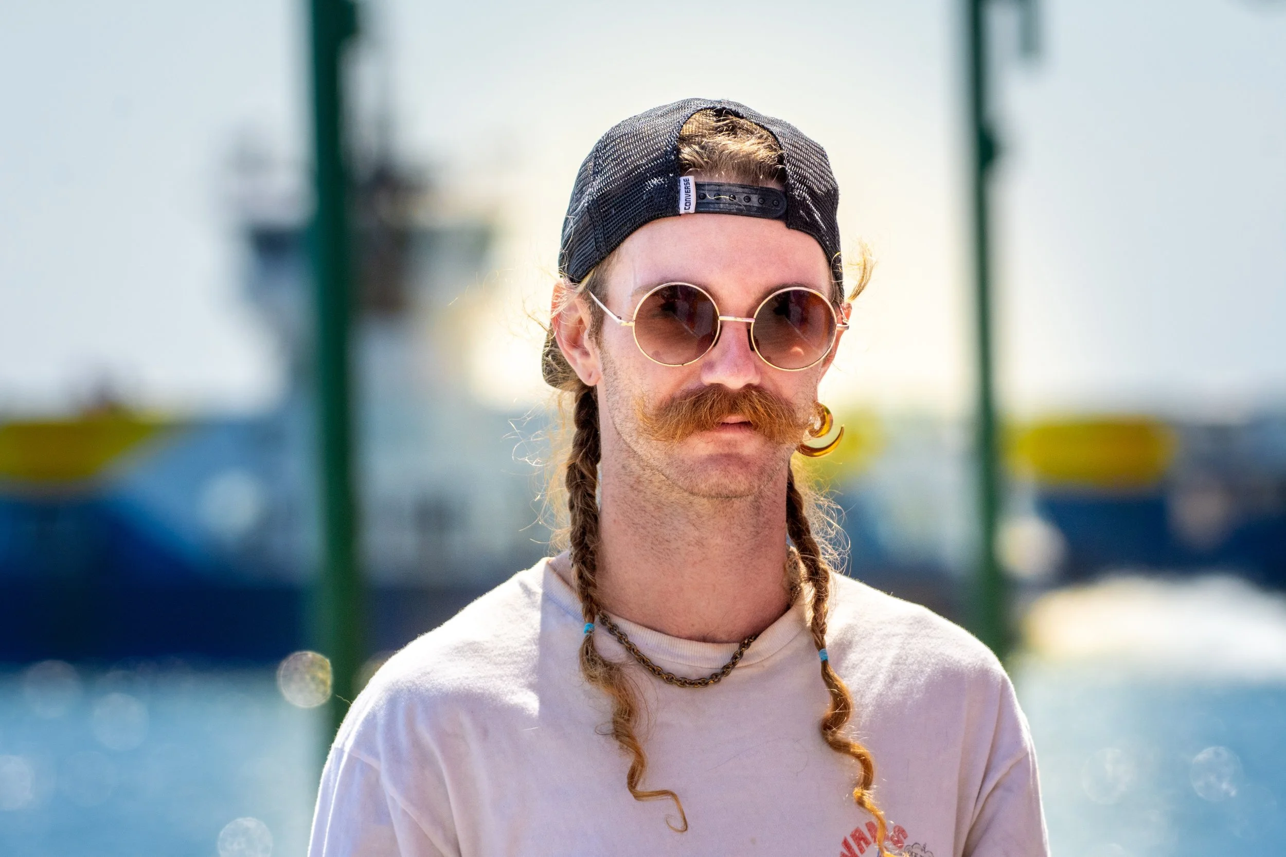 A man with a long braided hairstyle, wearing round sunglasses, a black snapback cap, and a white T-shirt with red writing, standing outdoors with a blurred background of green and blue.