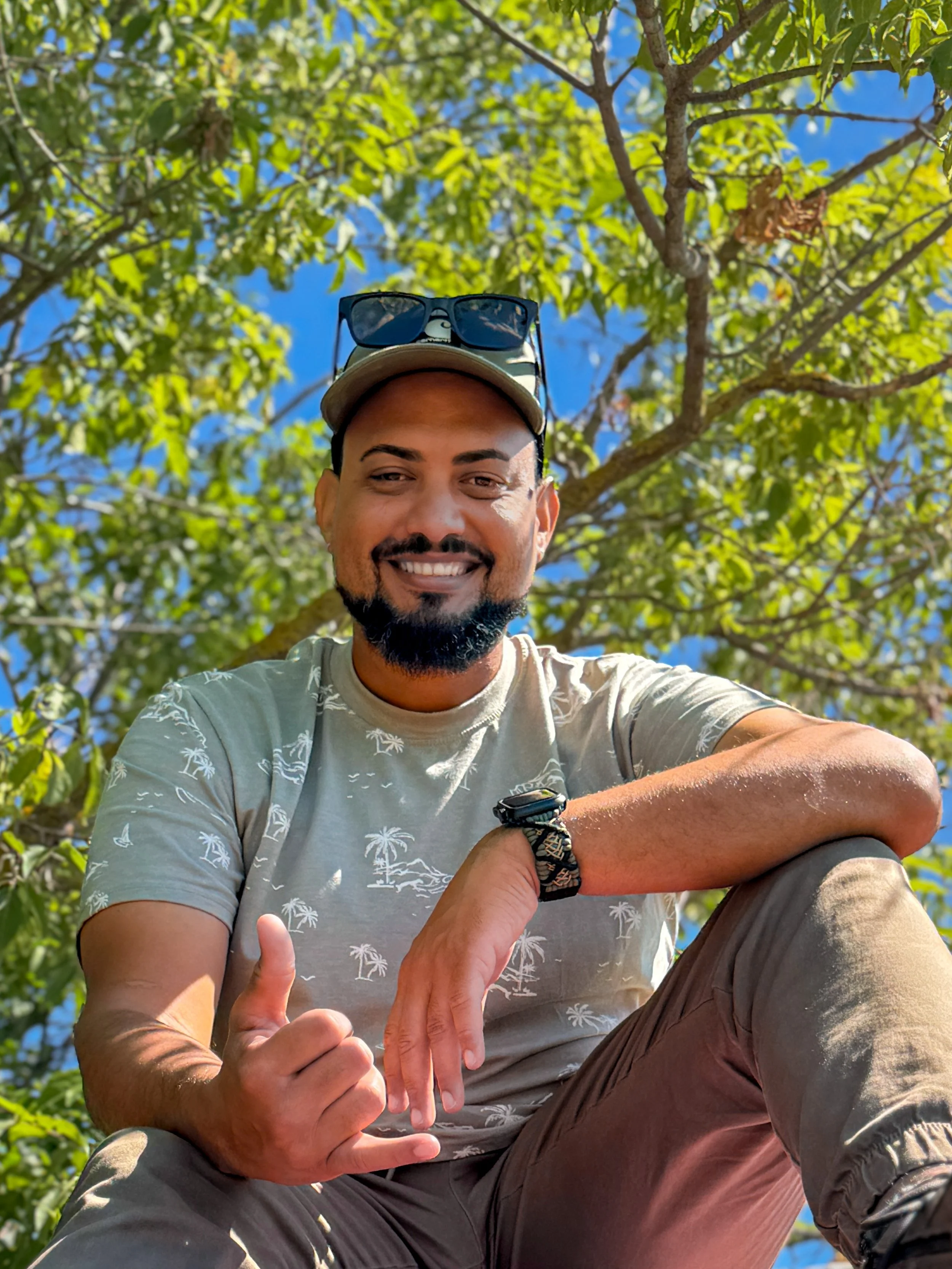 A smiling man wearing sunglasses on his cap, sitting outdoors under a tree with green leaves and a bright blue sky, giving a thumbs-up.