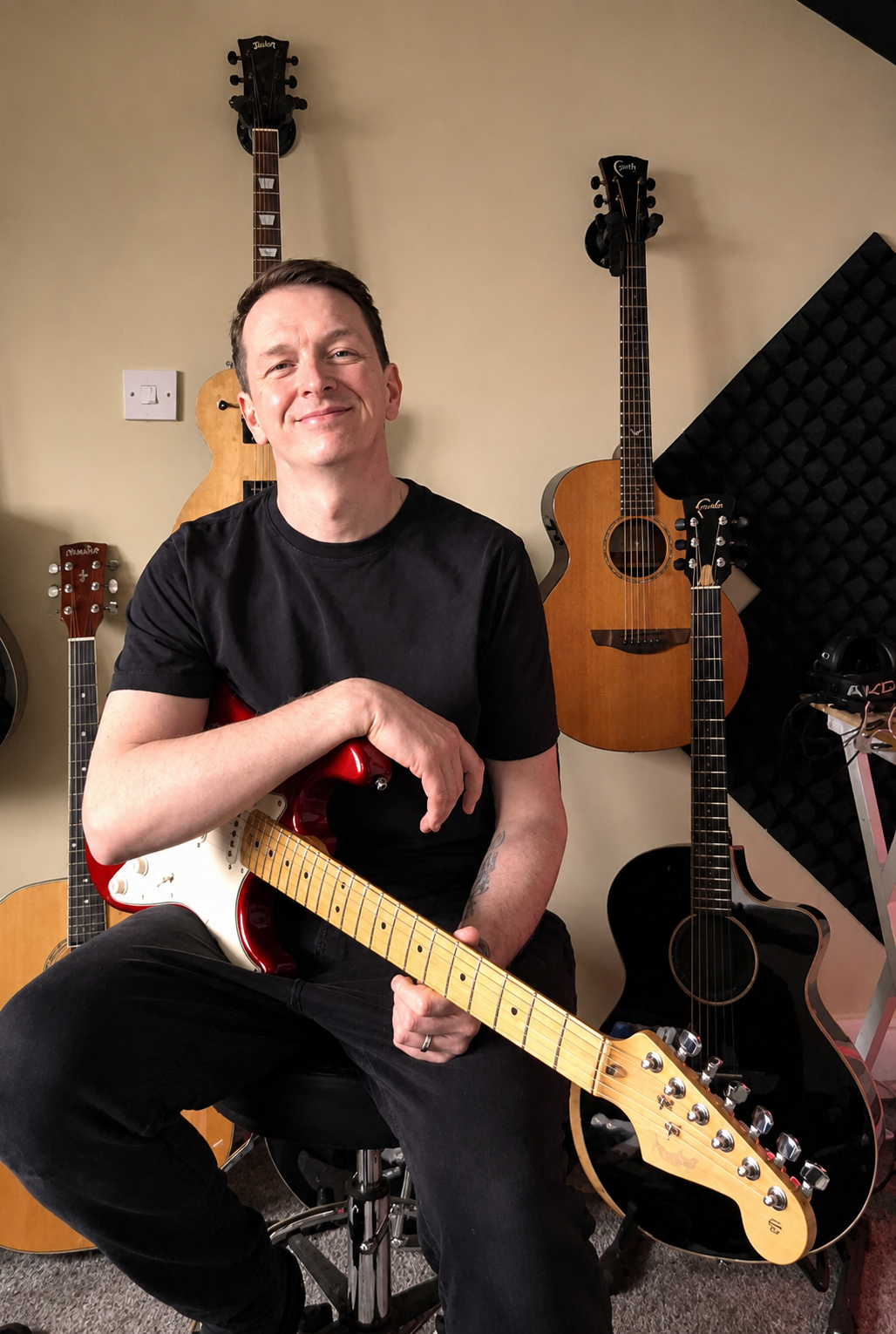 A man sitting on a stool holding a red electric guitar, surrounded by acoustic guitars hanging on the wall and resting on the floor in a music studio.