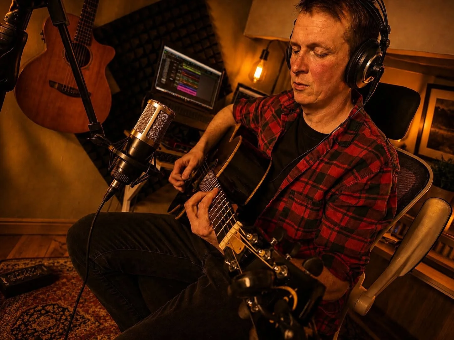 A man wearing a red plaid shirt and headphones playing an acoustic guitar in a recording studio.